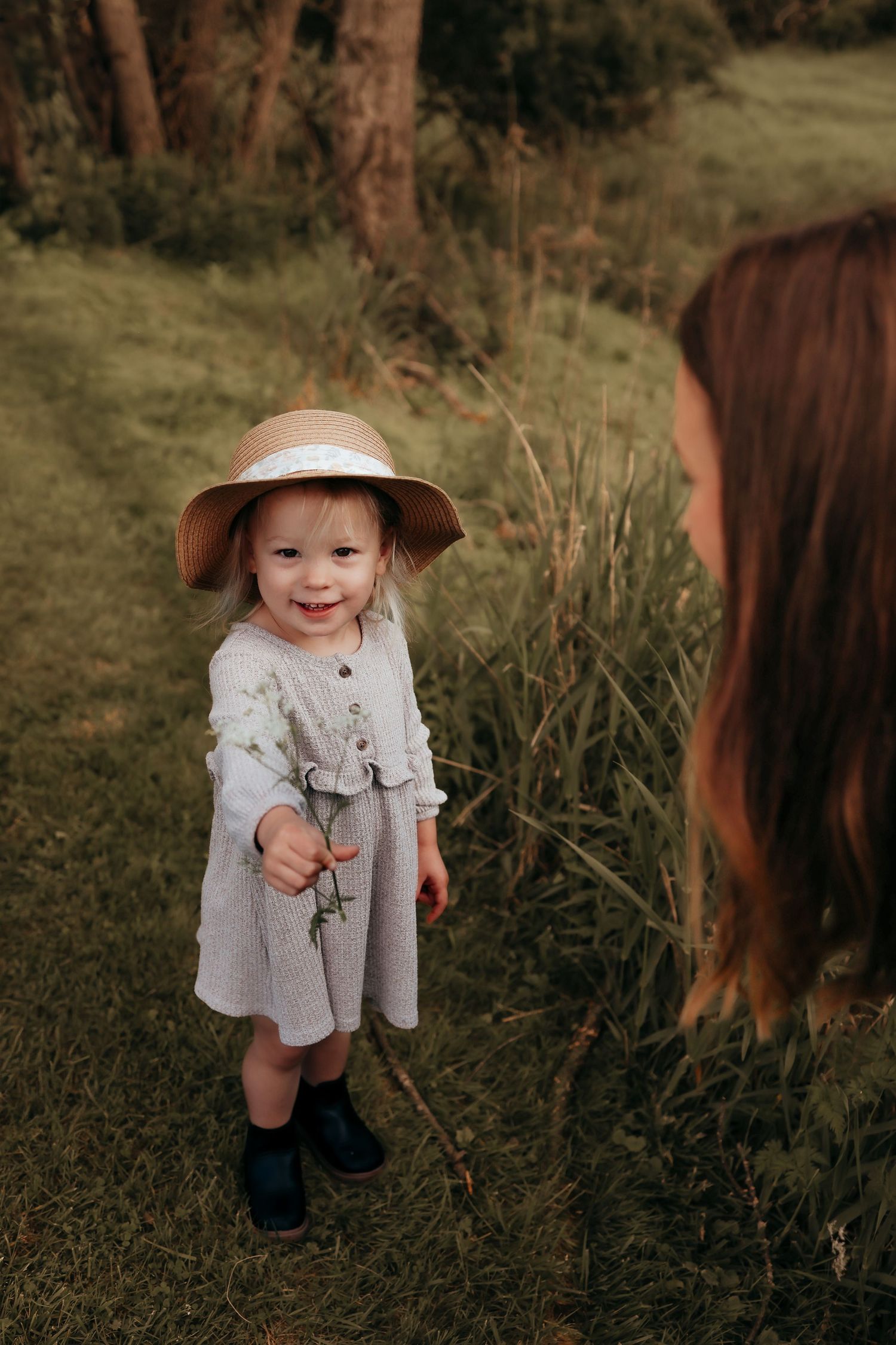 Smiling toddler girl in straw hat and gray dress holds a flower in a lush green outdoor setting.