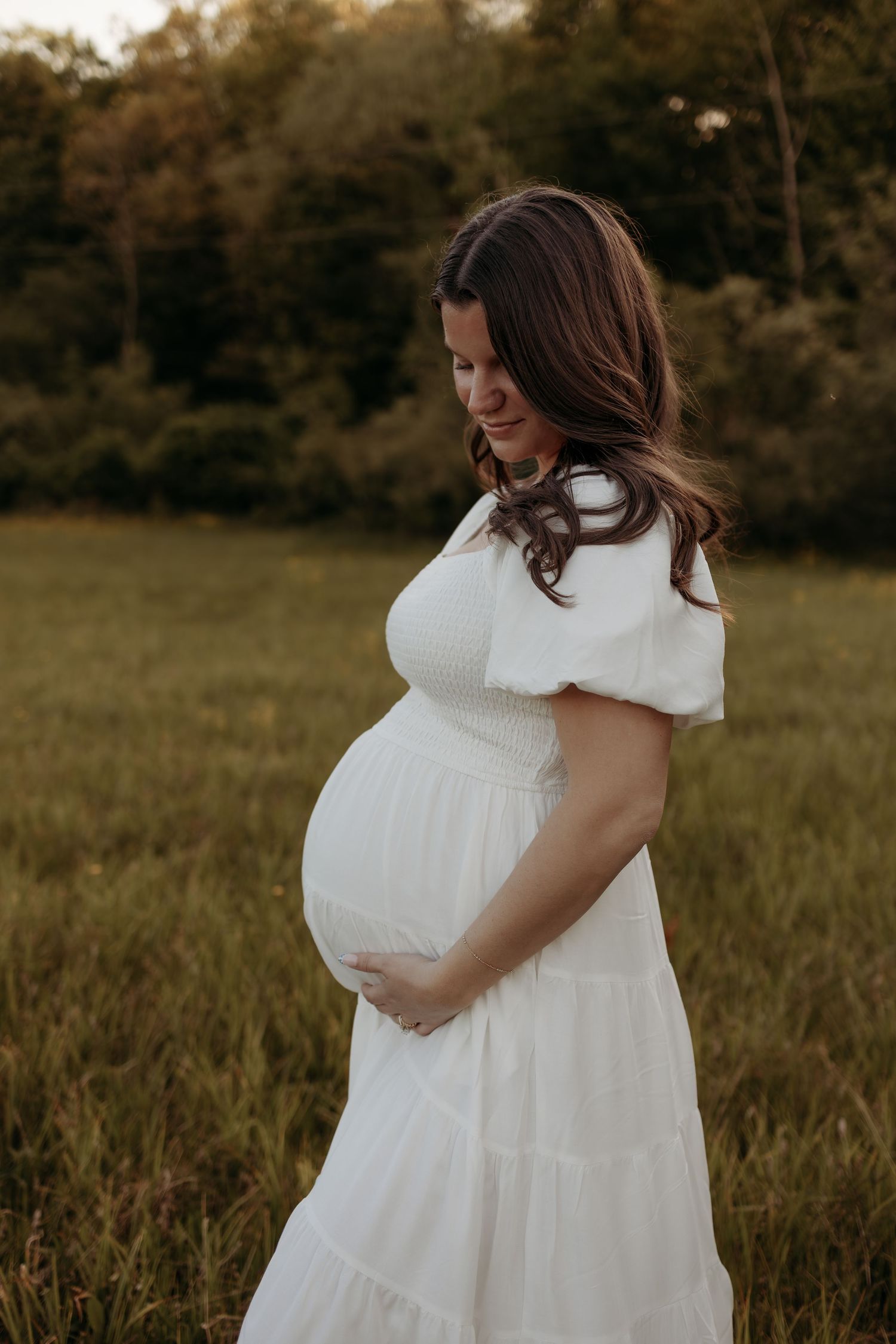 Pregnant woman in white dress cradling baby bump in golden hour field.