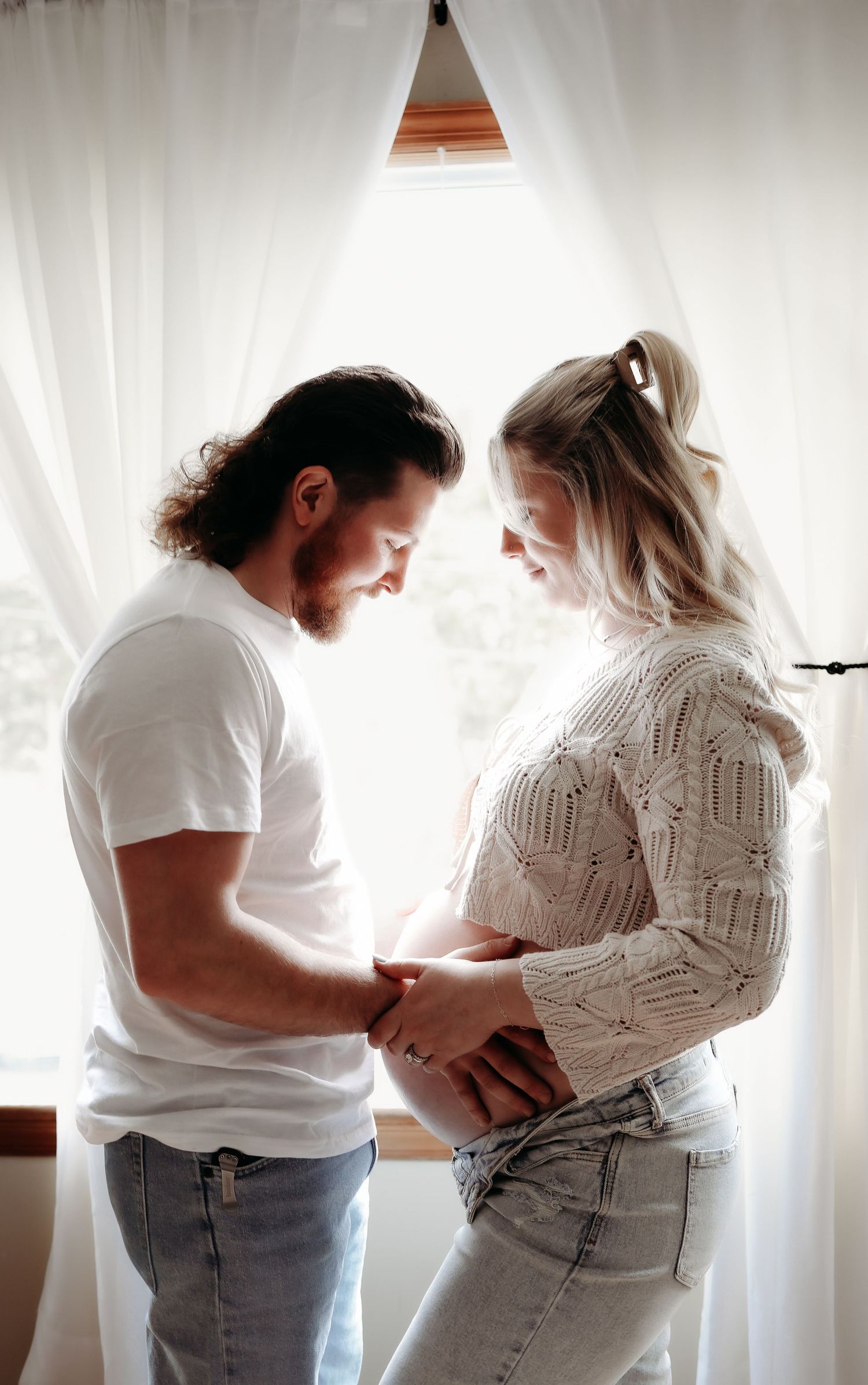 Couple tenderly holding pregnant belly together while standing by a bright window.
