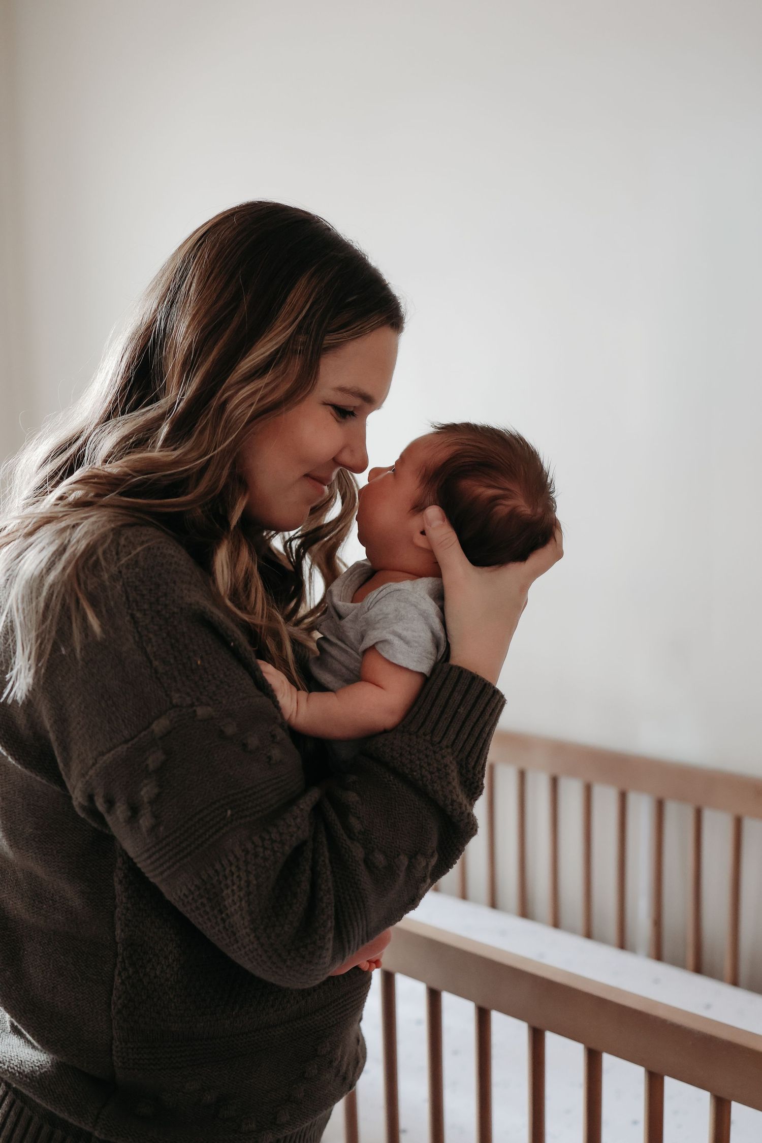 Mother tenderly touching noses with newborn baby while standing beside wooden crib.