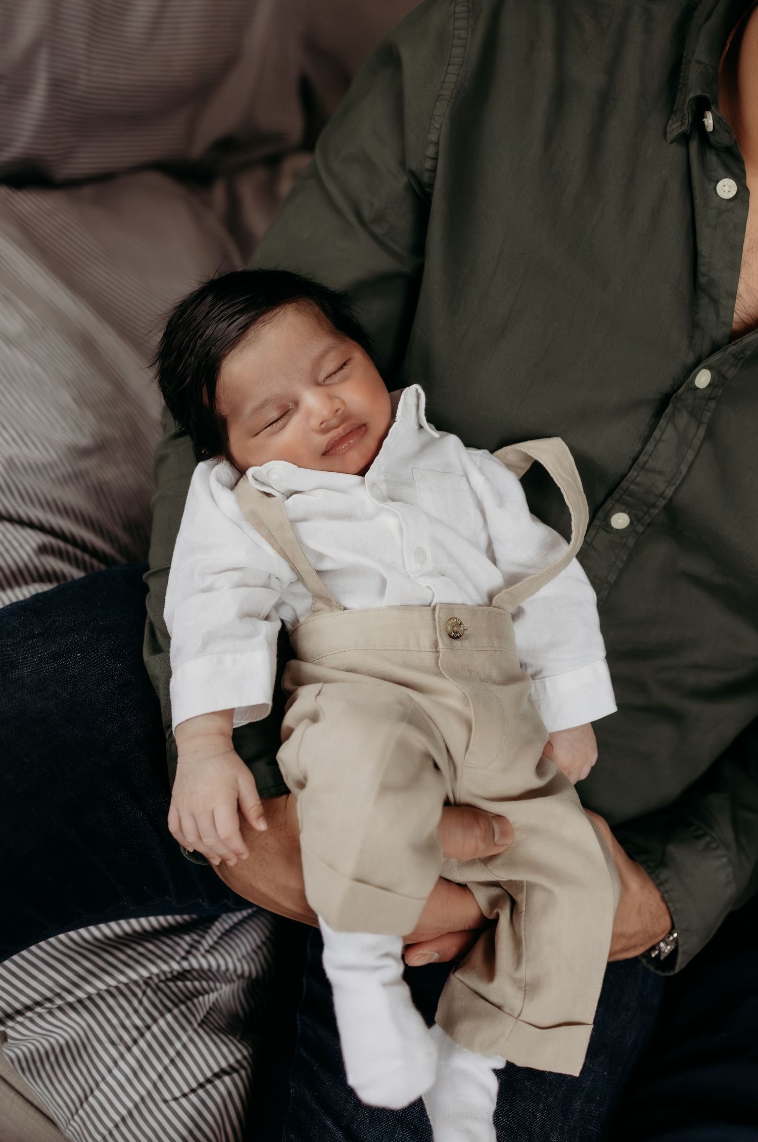 A peacefully sleeping newborn in white shirt and tan suspender pants cradled in parents arms.