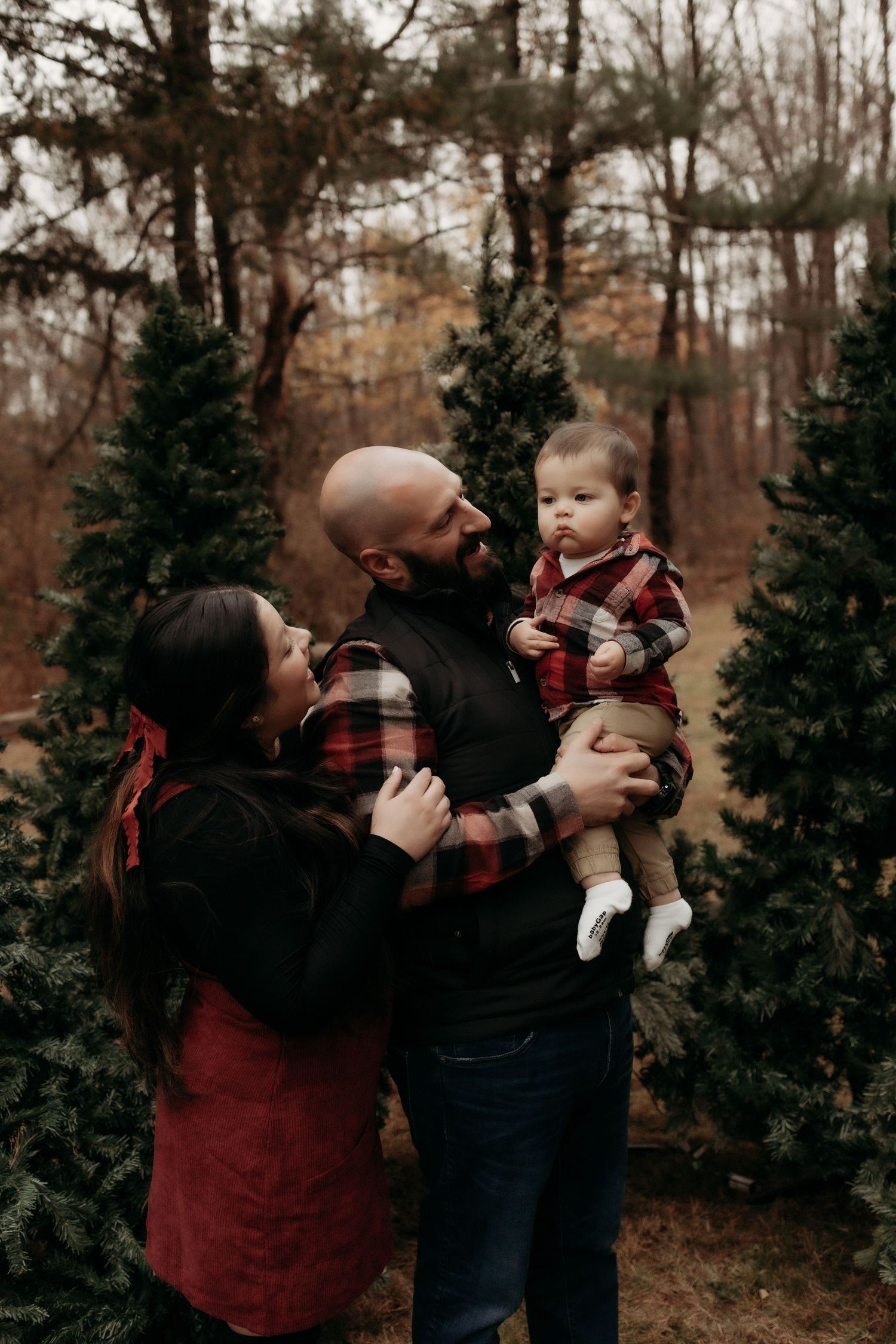 Family of three poses among evergreen trees, parents in red plaid holding toddler during a festive Christmas tree farm photo session.