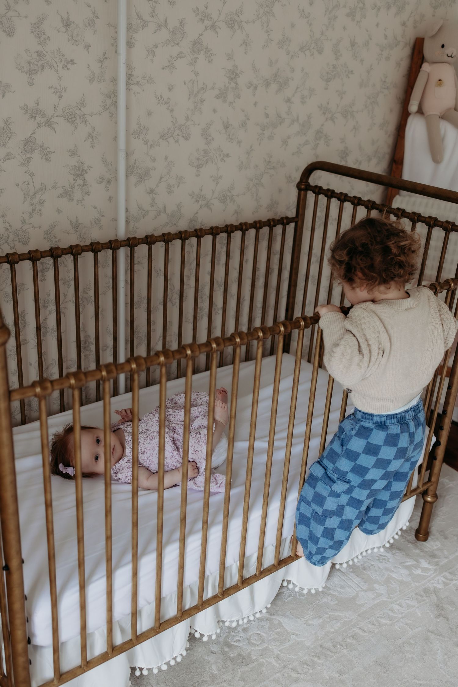 Curious toddler in checkered pants peering into vintage brass crib at newborn baby in floral nursery.
