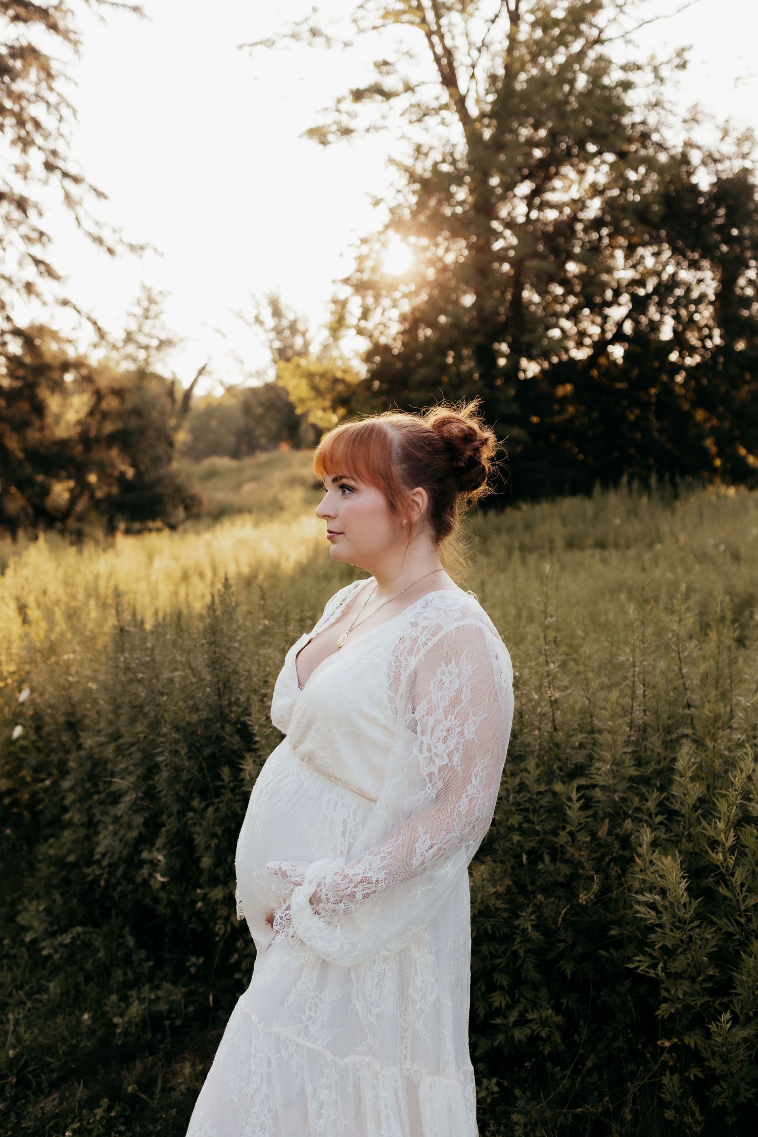 Pregnant woman in white lace dress standing in golden hour sunlight among green meadow grasses.