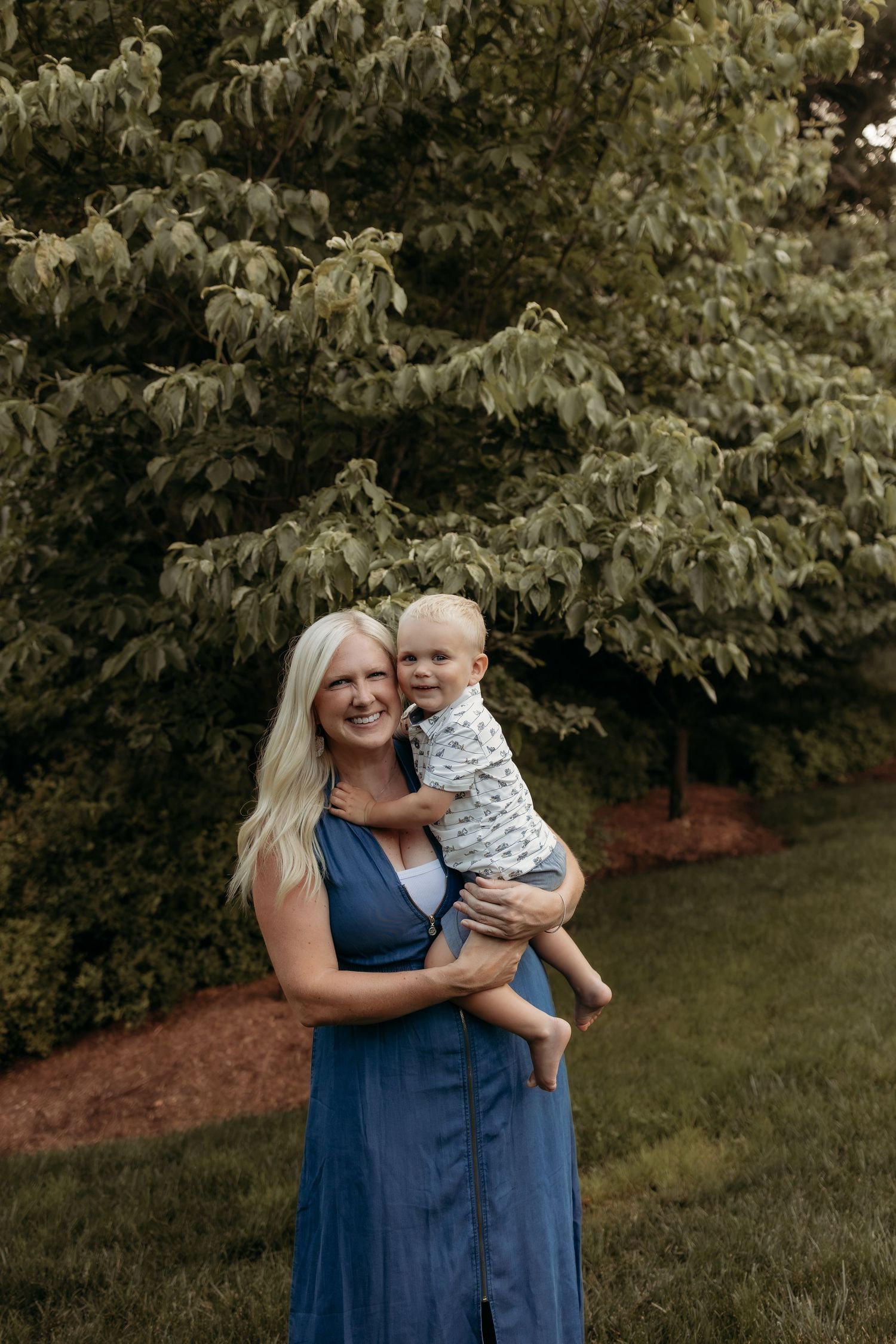 Smiling blonde woman in blue dress holding a happy toddler outdoors with lush green trees in background.
