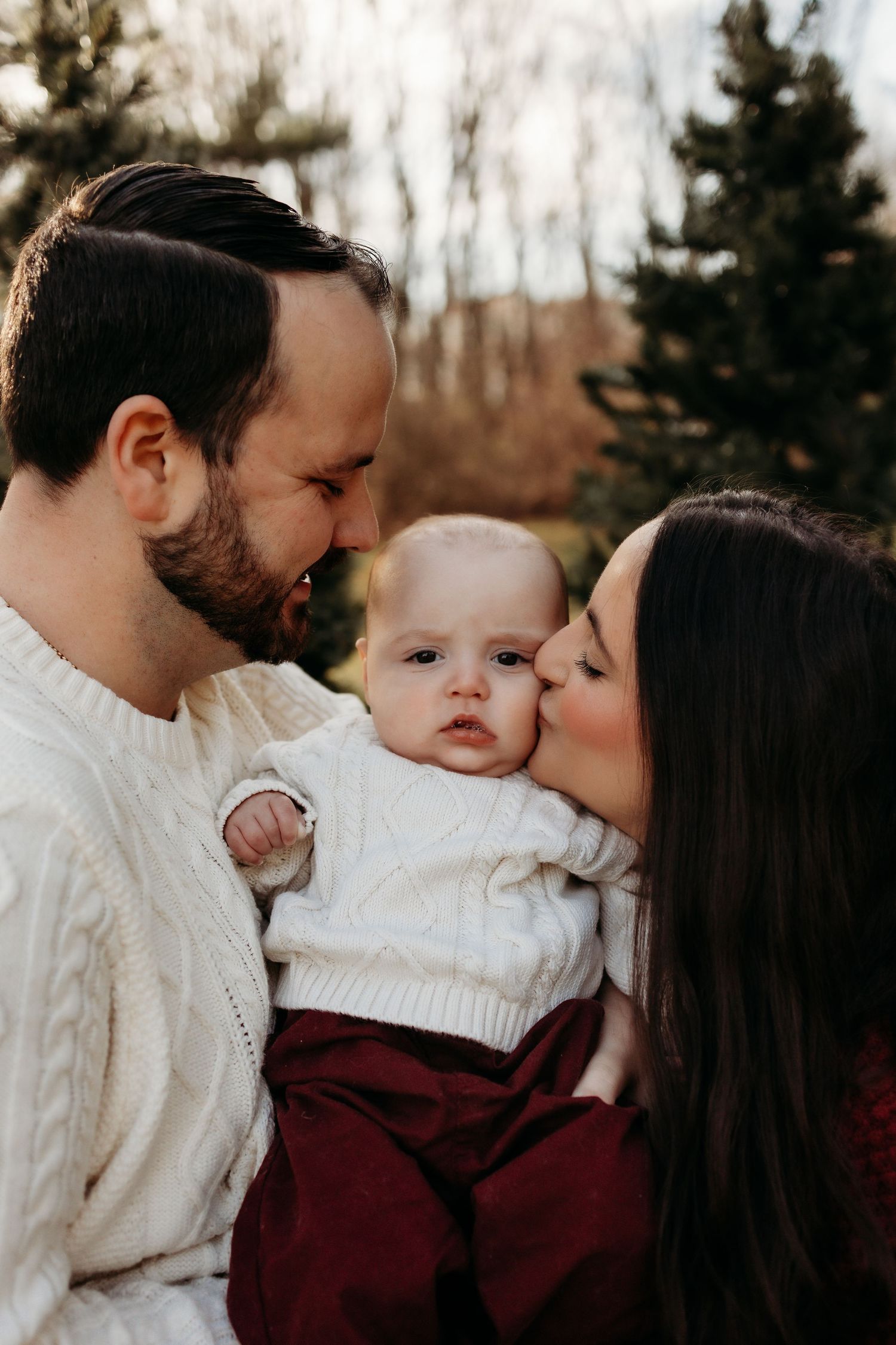 A couple in white cable-knit sweaters lovingly kiss their baby outdoors with evergreen trees in the background.