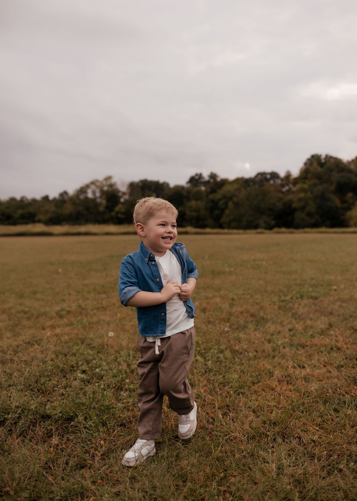 A happy toddler boy in a blue shirt and brown pants laughs while standing in a green open field.