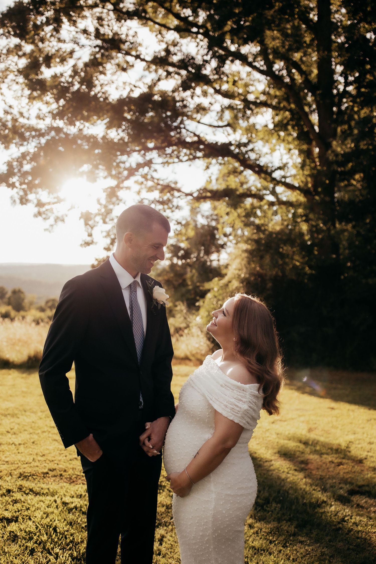 A couple in wedding attire holds hands at golden hour, smiling at each other in a sunlit outdoor setting.