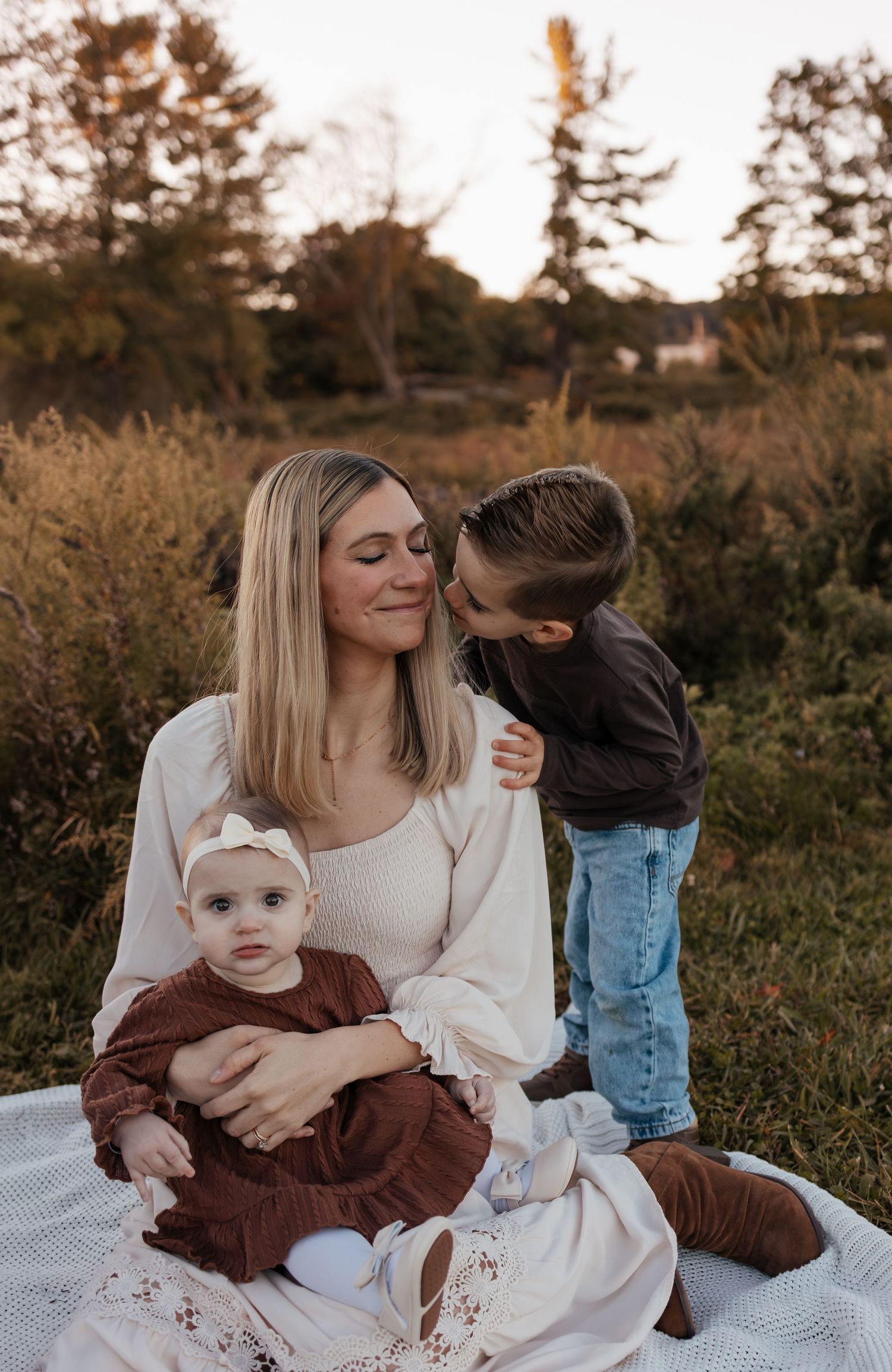 A smiling mother sits on a blanket in a golden autumn field holding her baby while her young son leans in to kiss her cheek.