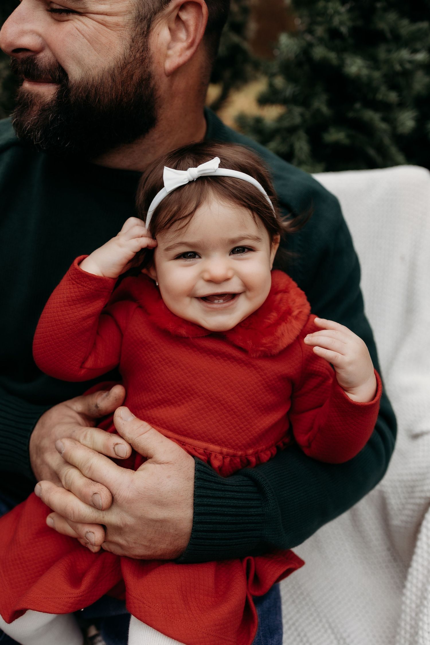 Smiling baby girl in red dress and white bow headband being held by bearded man in green sweater outdoors.