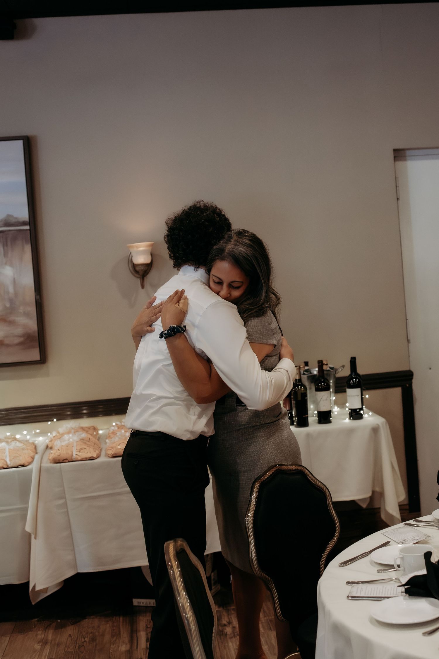 Two women sharing warm embrace at wedding reception dinner in elegant restaurant setting.
