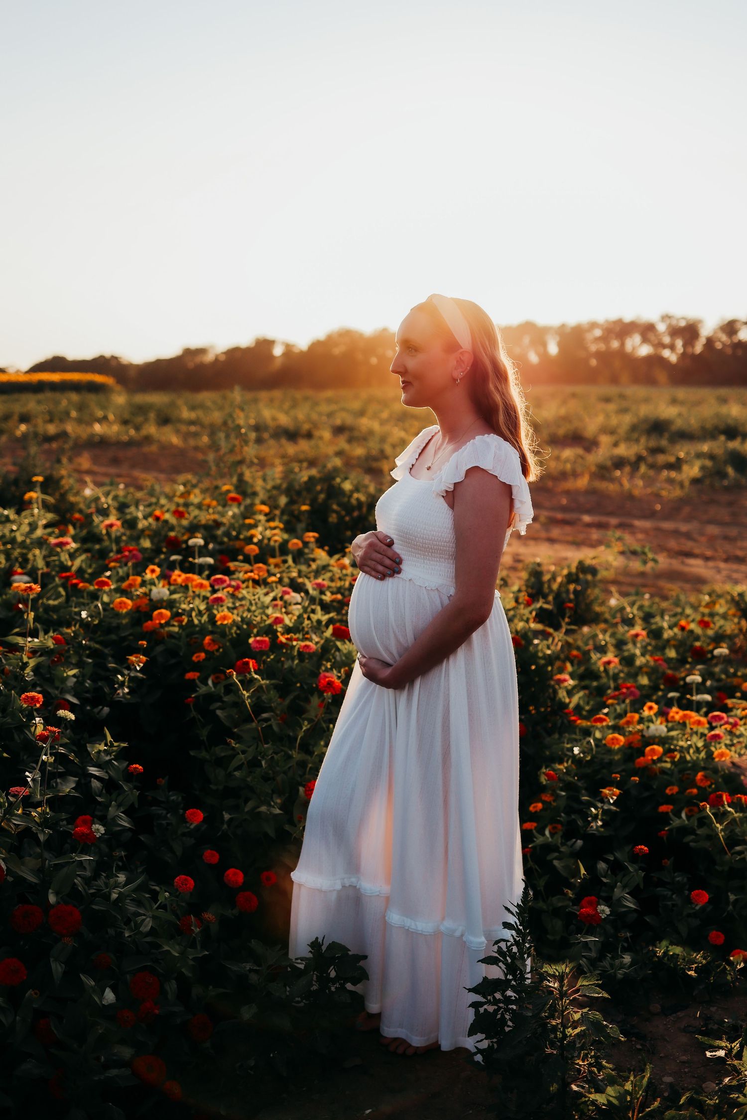 A pregnant woman in a flowing white dress stands in a golden-lit flower field at sunset amidst orange blooms.