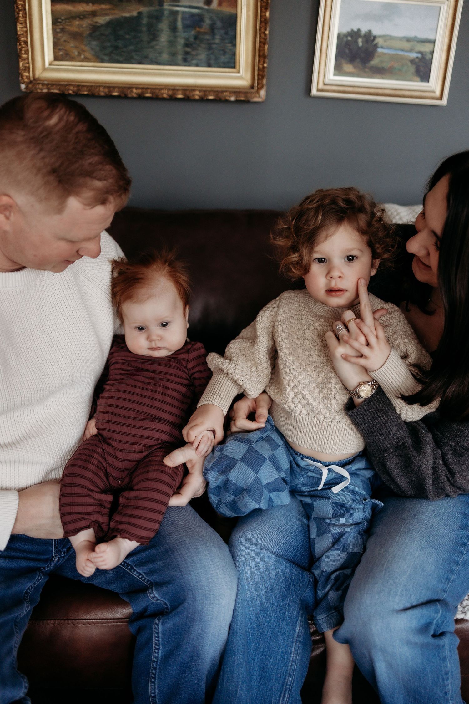 A family of four with two red-haired toddlers sits together on a leather couch at home.