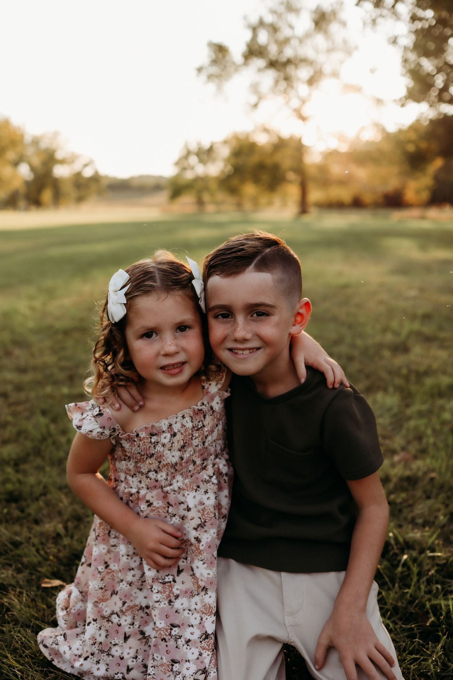 A smiling young boy and girl in a floral dress pose together at golden hour outdoors.