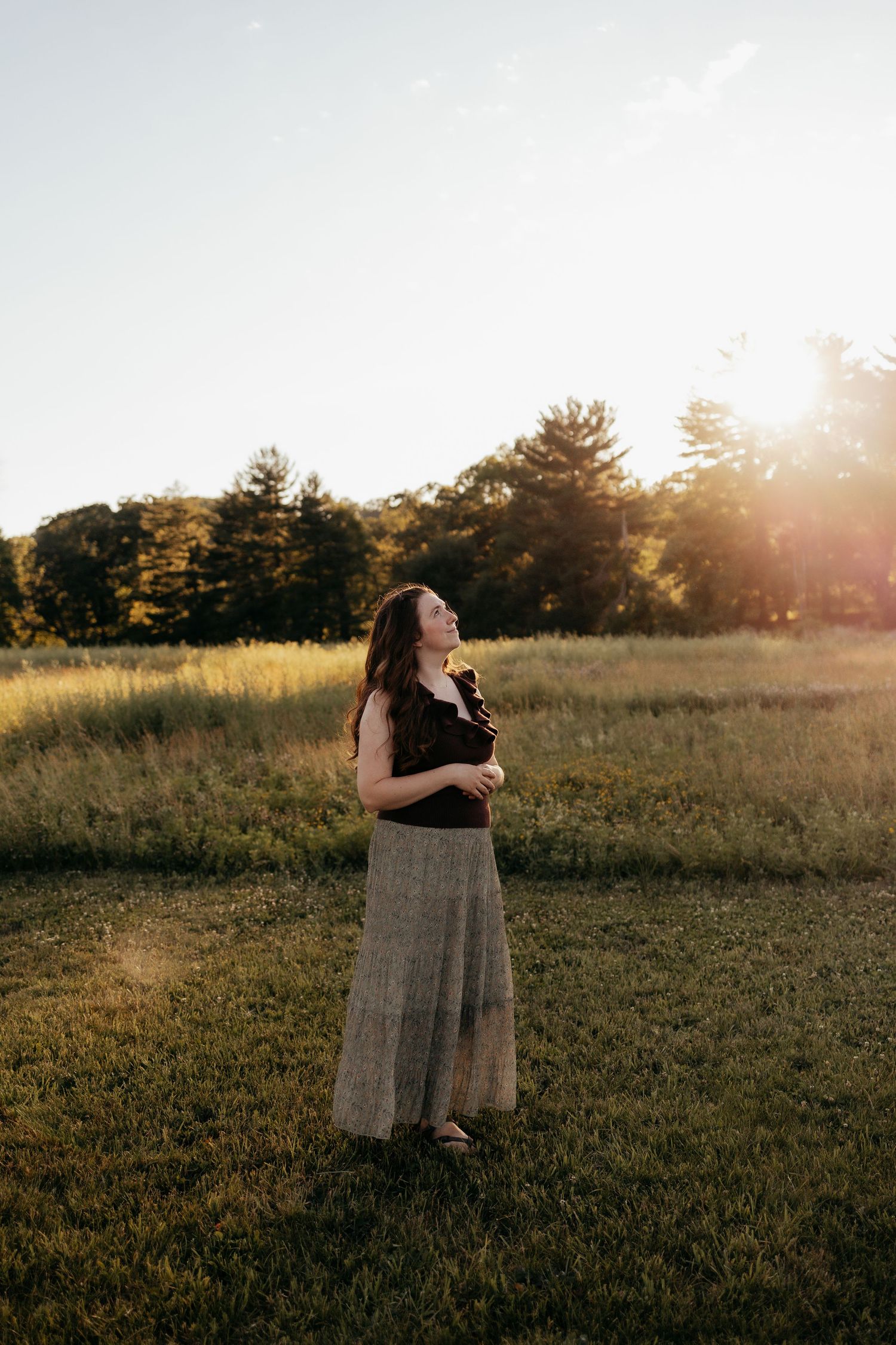 Woman in black top and maxi skirt gazes upward in golden meadow with warm sunset light glowing behind trees.
