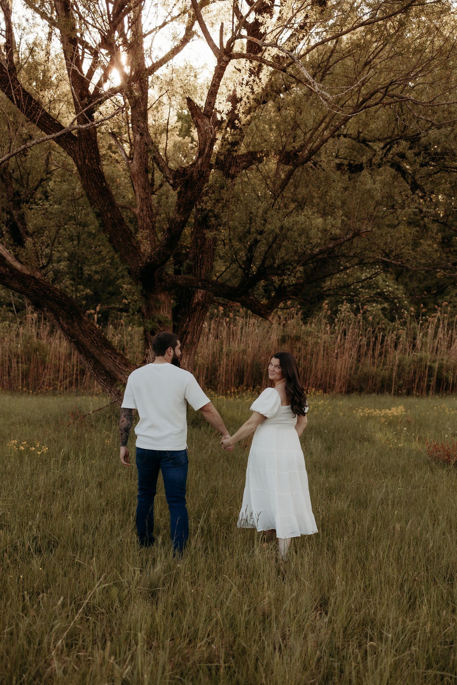 Couple holding hands walking toward large tree in golden hour field.