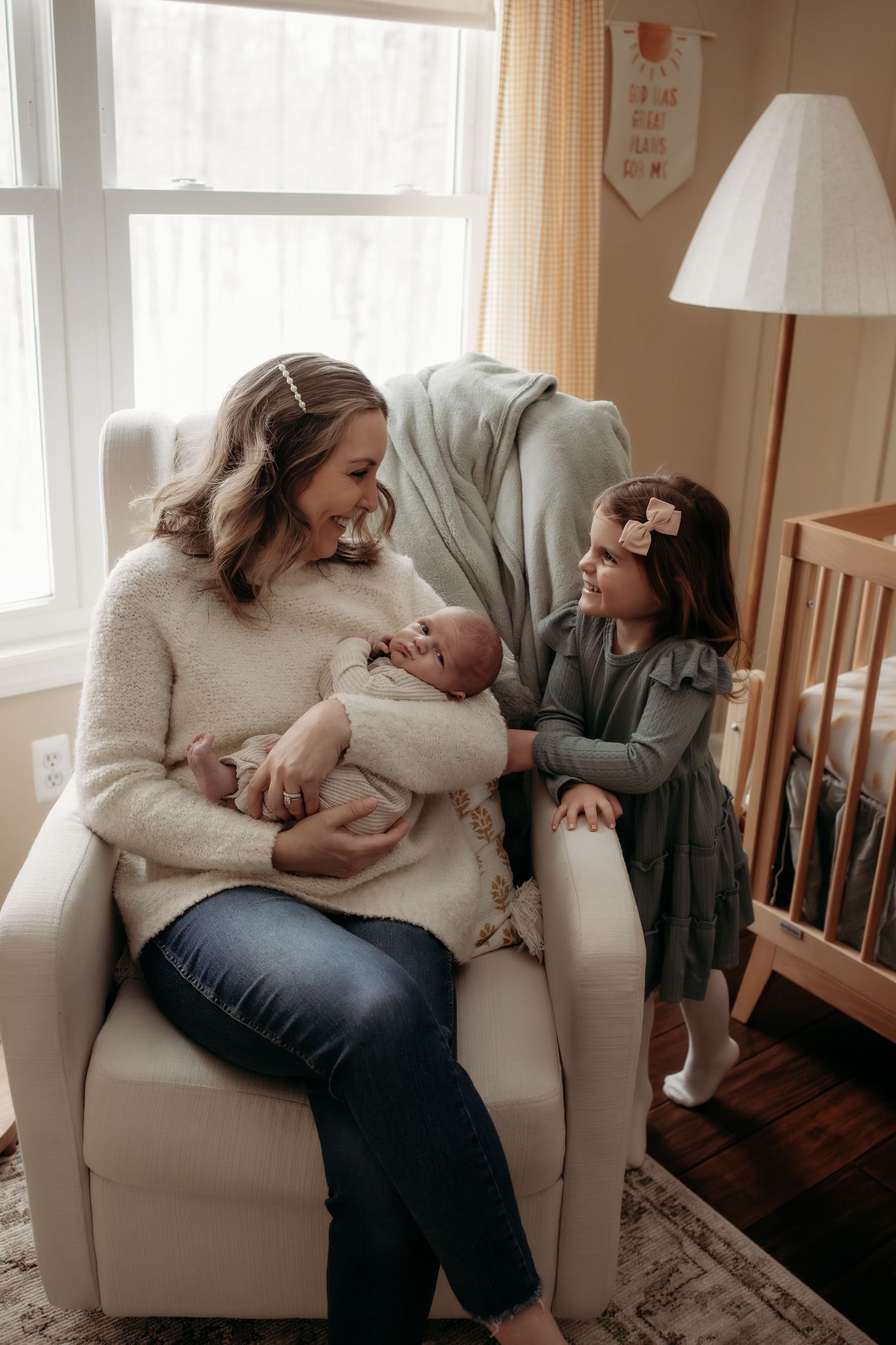 Mother holds newborn while young toddler daughter in gray dress reaches out in nursery chair.