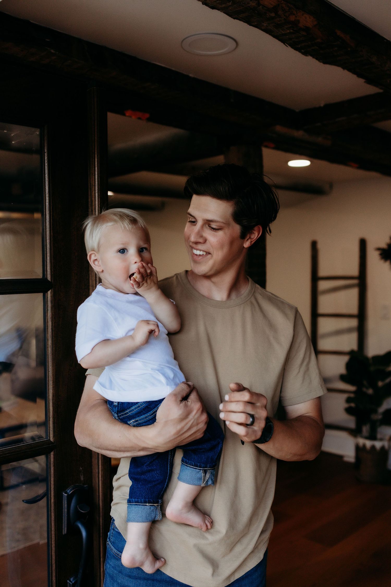 Young man in tan shirt smiling while holding a happy toddler in a cozy indoor setting.