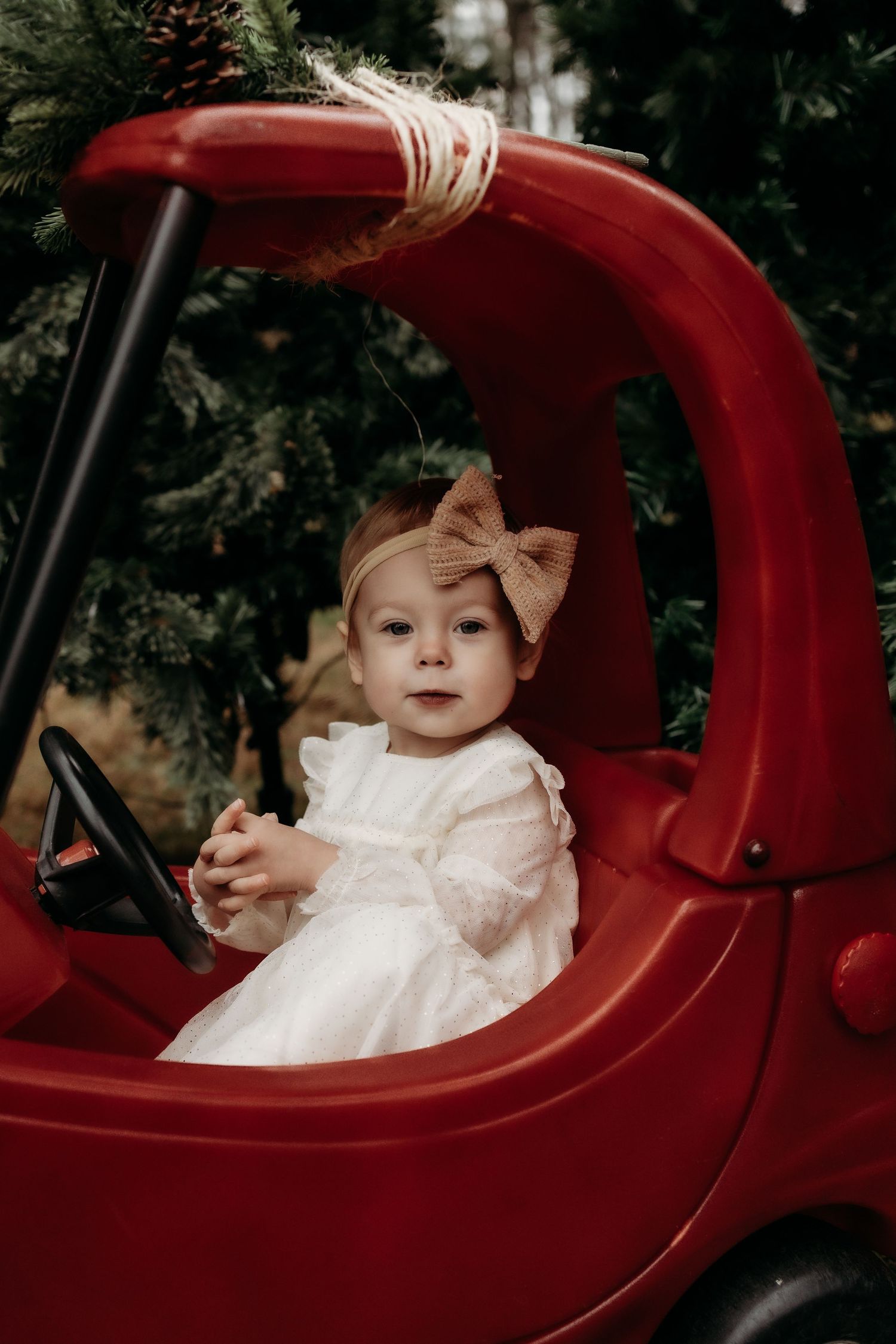 Toddler girl in white dress and bow headband sitting in red toy car surrounded by Christmas trees.