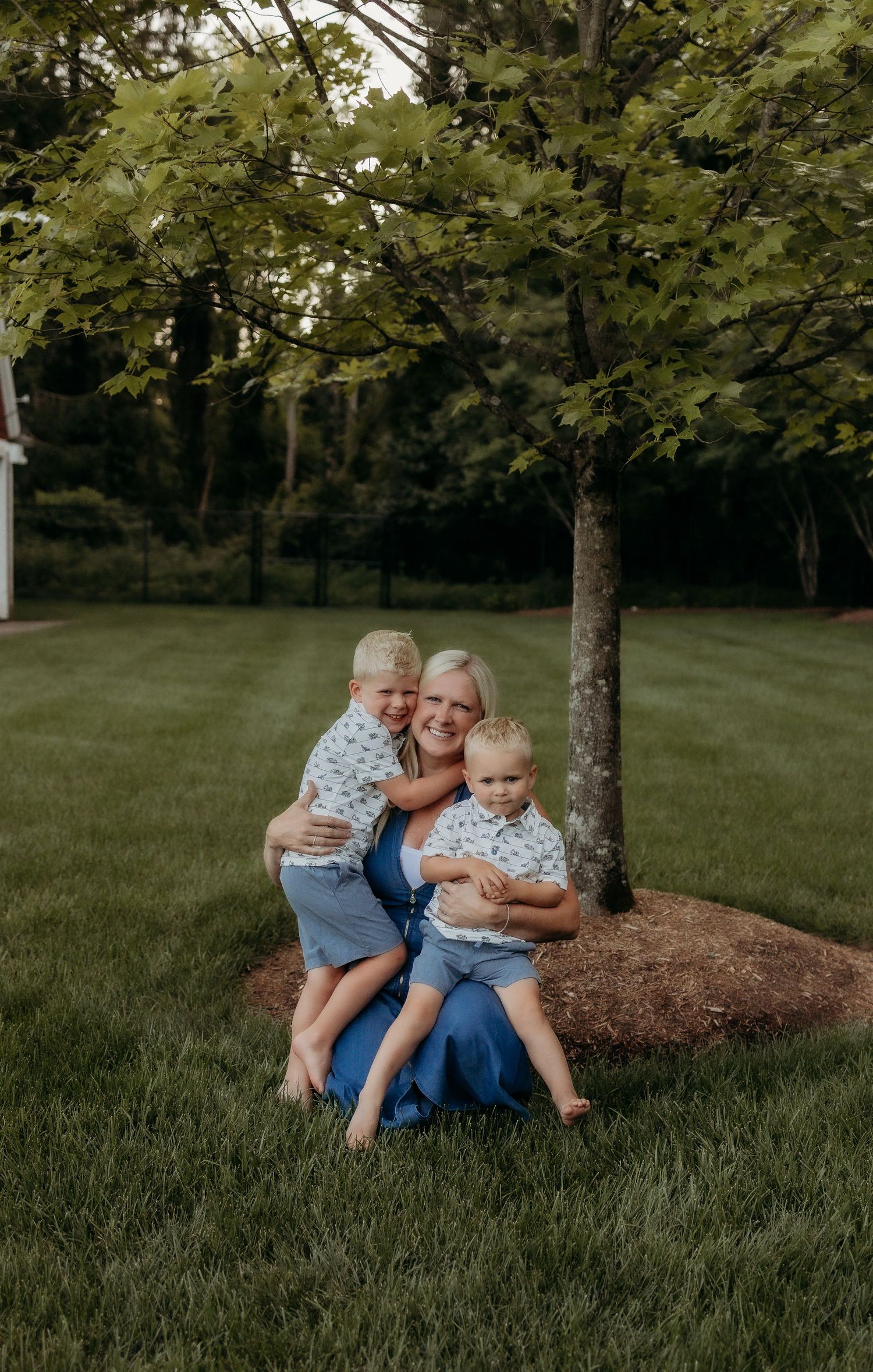 A smiling blonde woman sits on green grass hugging two young boys in matching floral shirts near a tree.