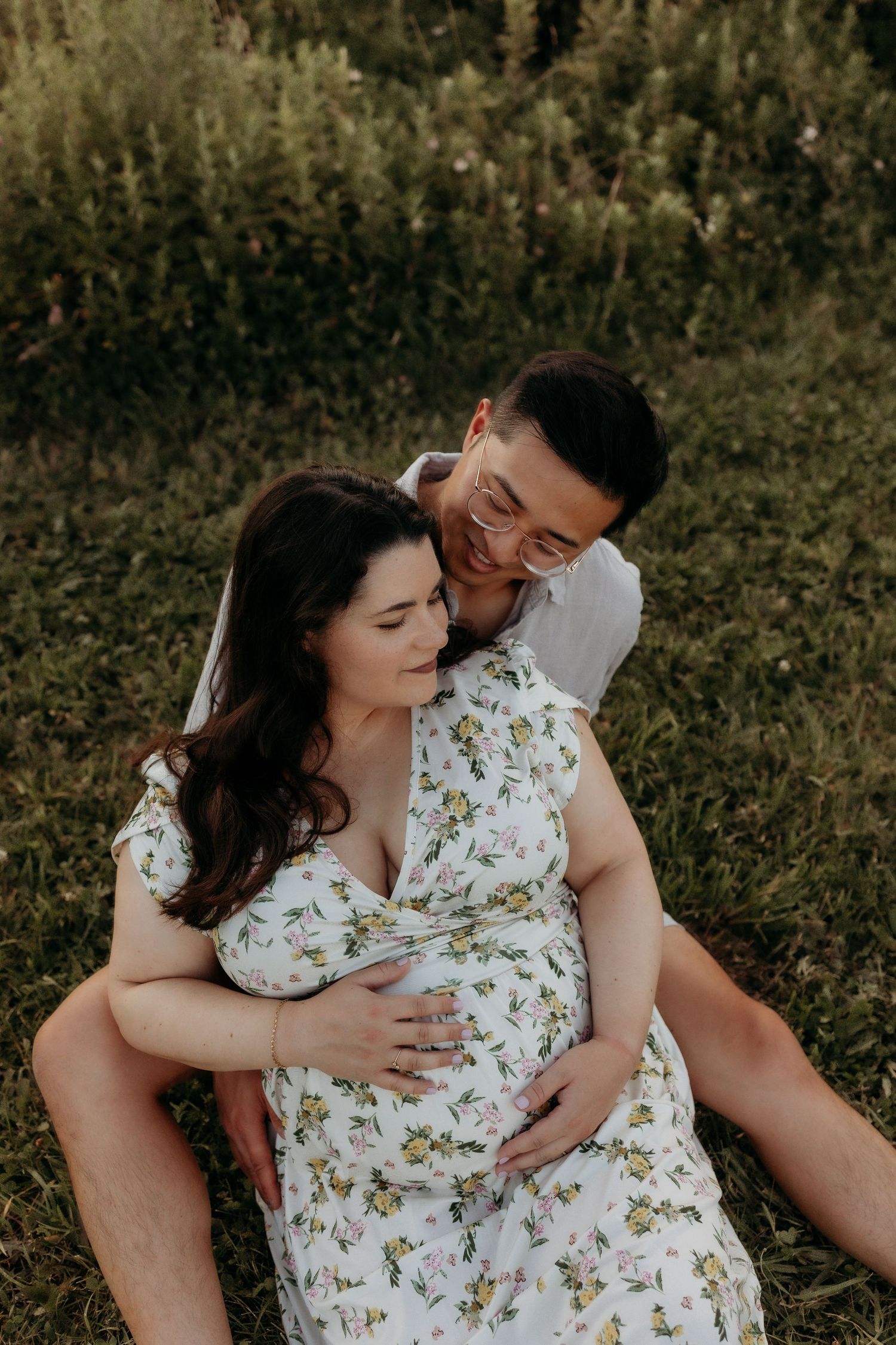 A smiling couple sits in a grassy field, man embracing pregnant woman in a floral dress.