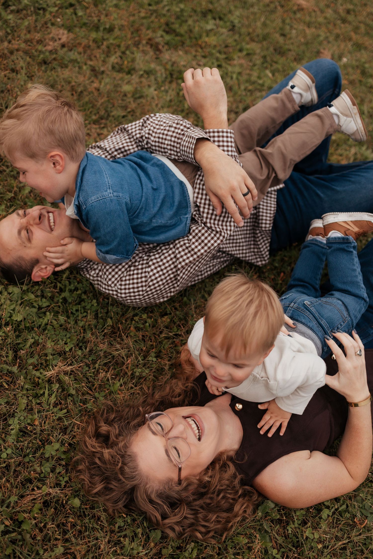Family of four laughing and playing together on green grass, two parents lying down with toddlers climbing on them.