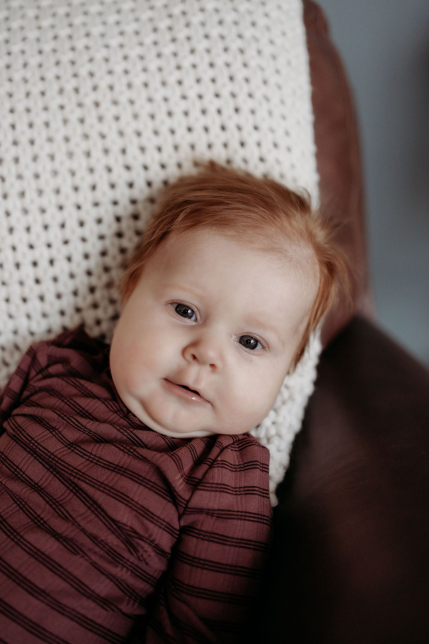 An adorable red-haired baby in a striped burgundy outfit rests against a white knit blanket.