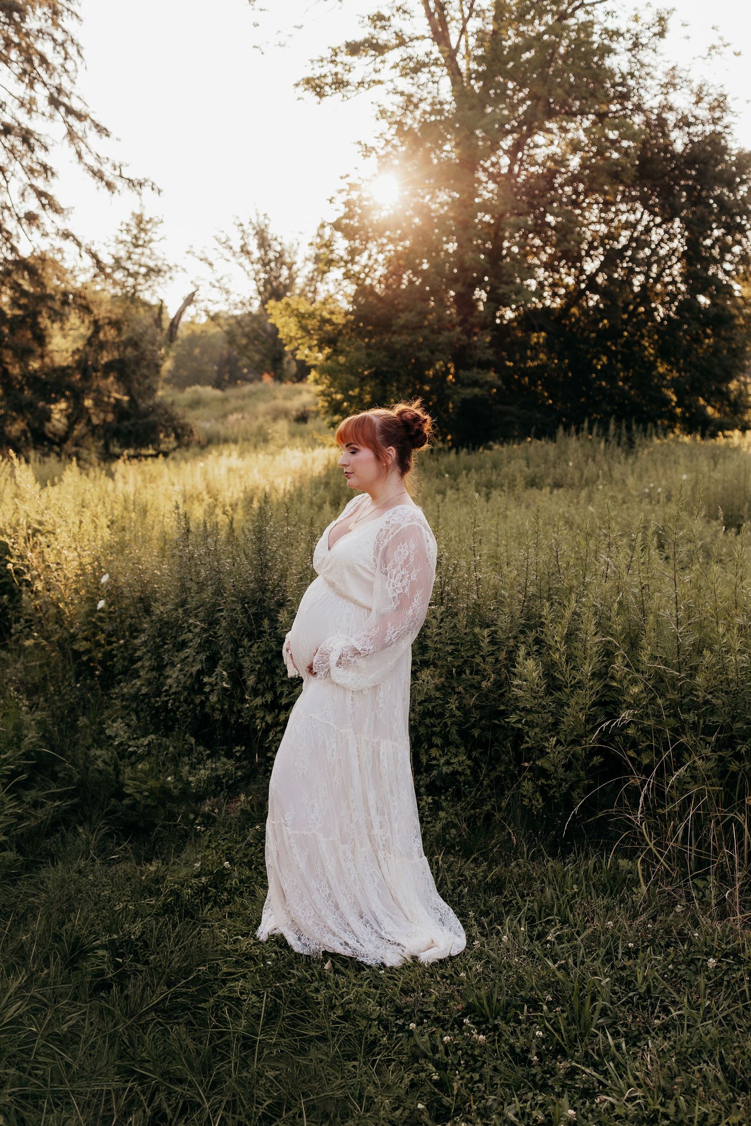 Pregnant woman in white lace dress standing in sunlit meadow during golden hour maternity photoshoot.