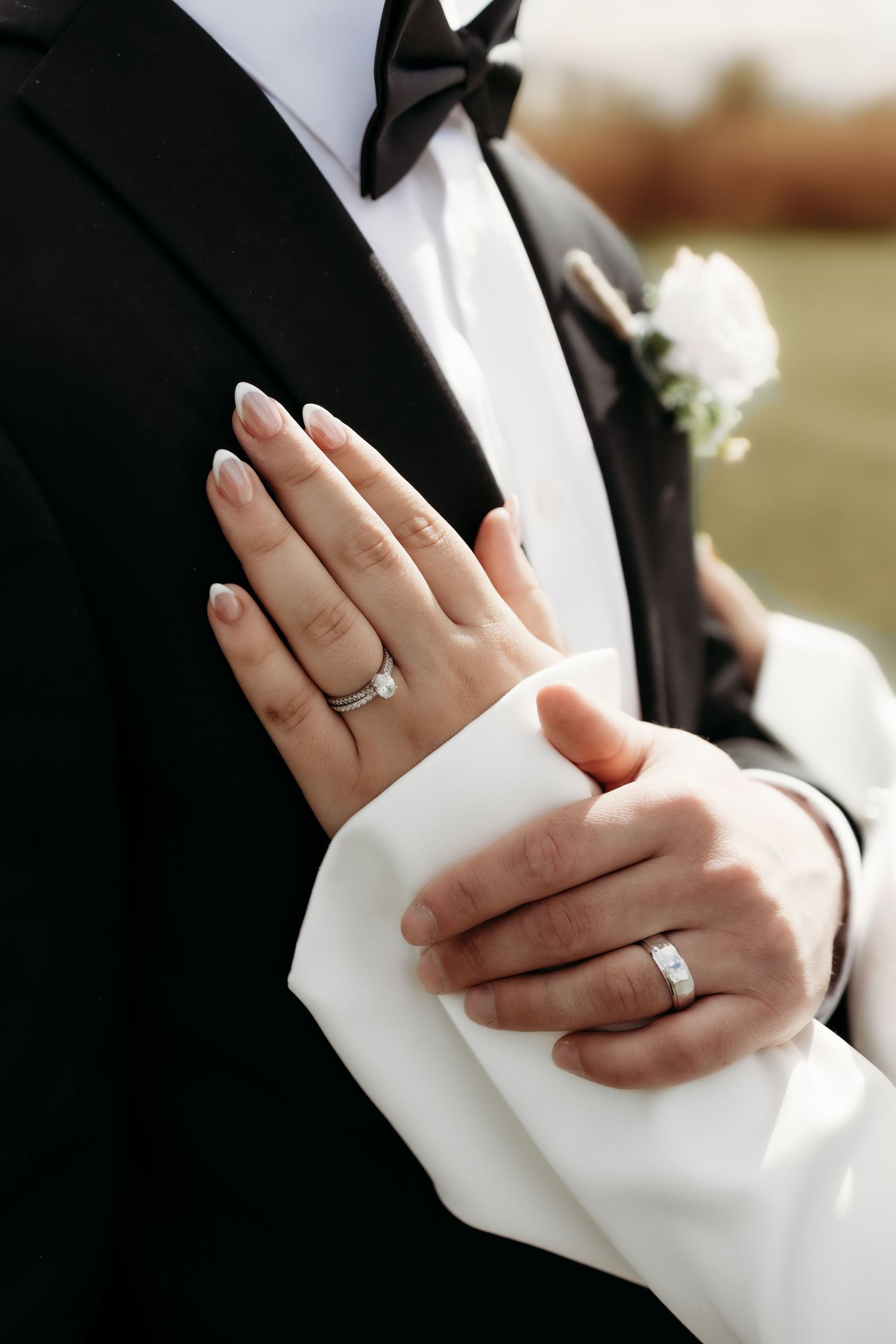 Close-up of bride and groom's hands showing wedding rings against groom's black tuxedo jacket.