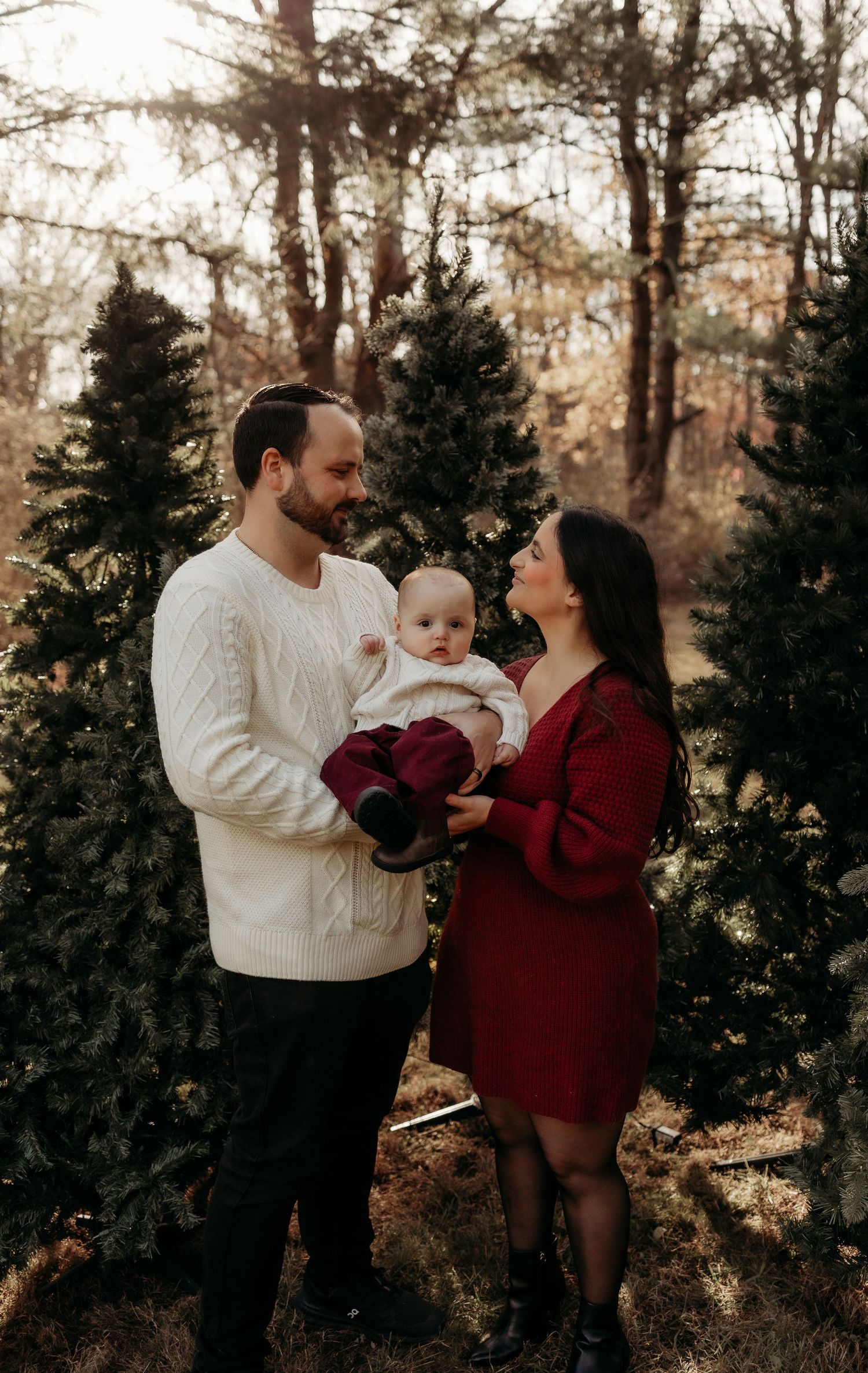 A young family poses among Christmas trees, father holding baby, mother in red dress, father in white sweater.