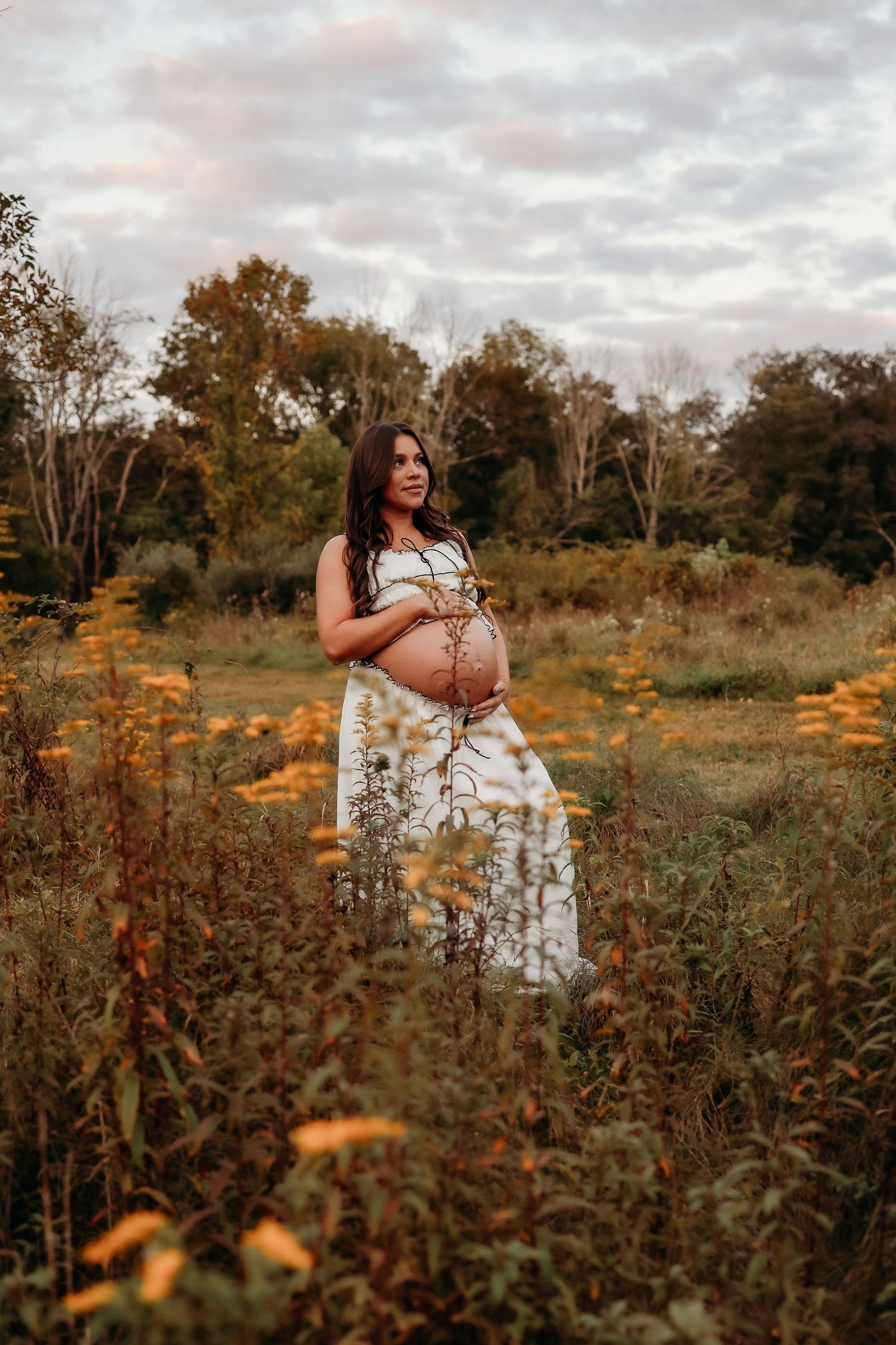 Pregnant woman in floral dress standing in an autumn wildflower field, gently holding her baby bump.