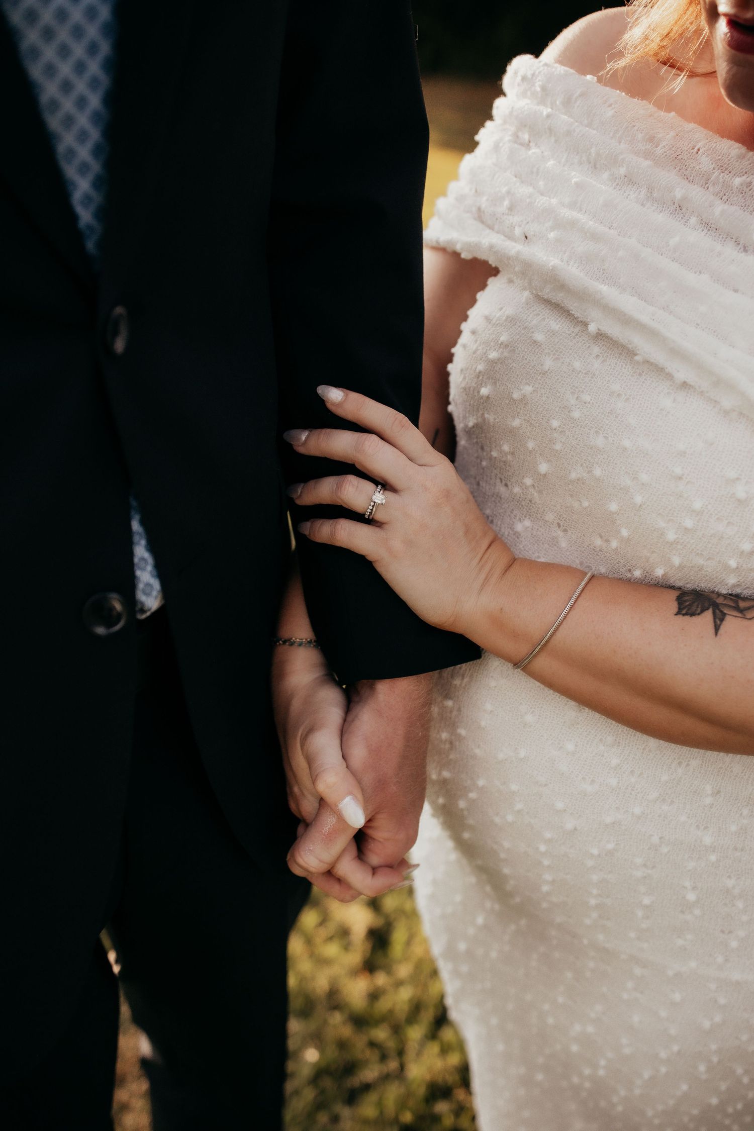 Bride and groom holding hands, showcasing wedding ring, tattoos, white off-shoulder dress and dark suit.