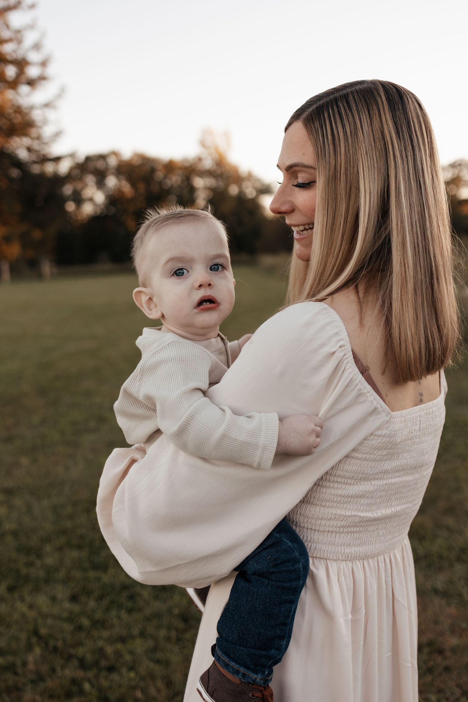 Smiling woman in a cream dress holding a wide-eyed baby in a neutral outfit while standing in a grassy park at golden hour.