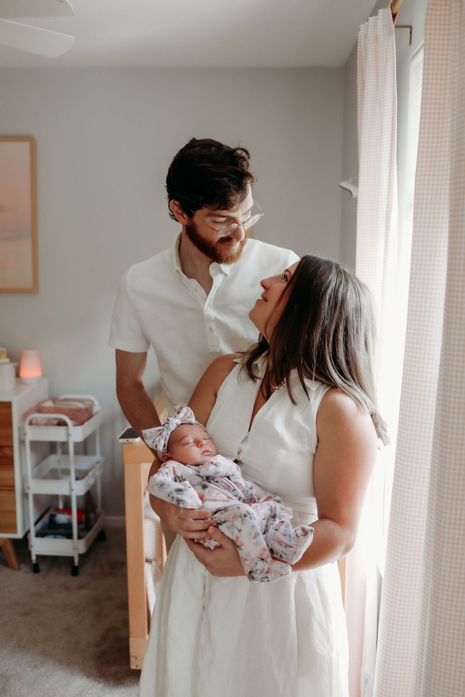 Young couple in white outfits sharing a smile while holding their swaddled newborn baby.