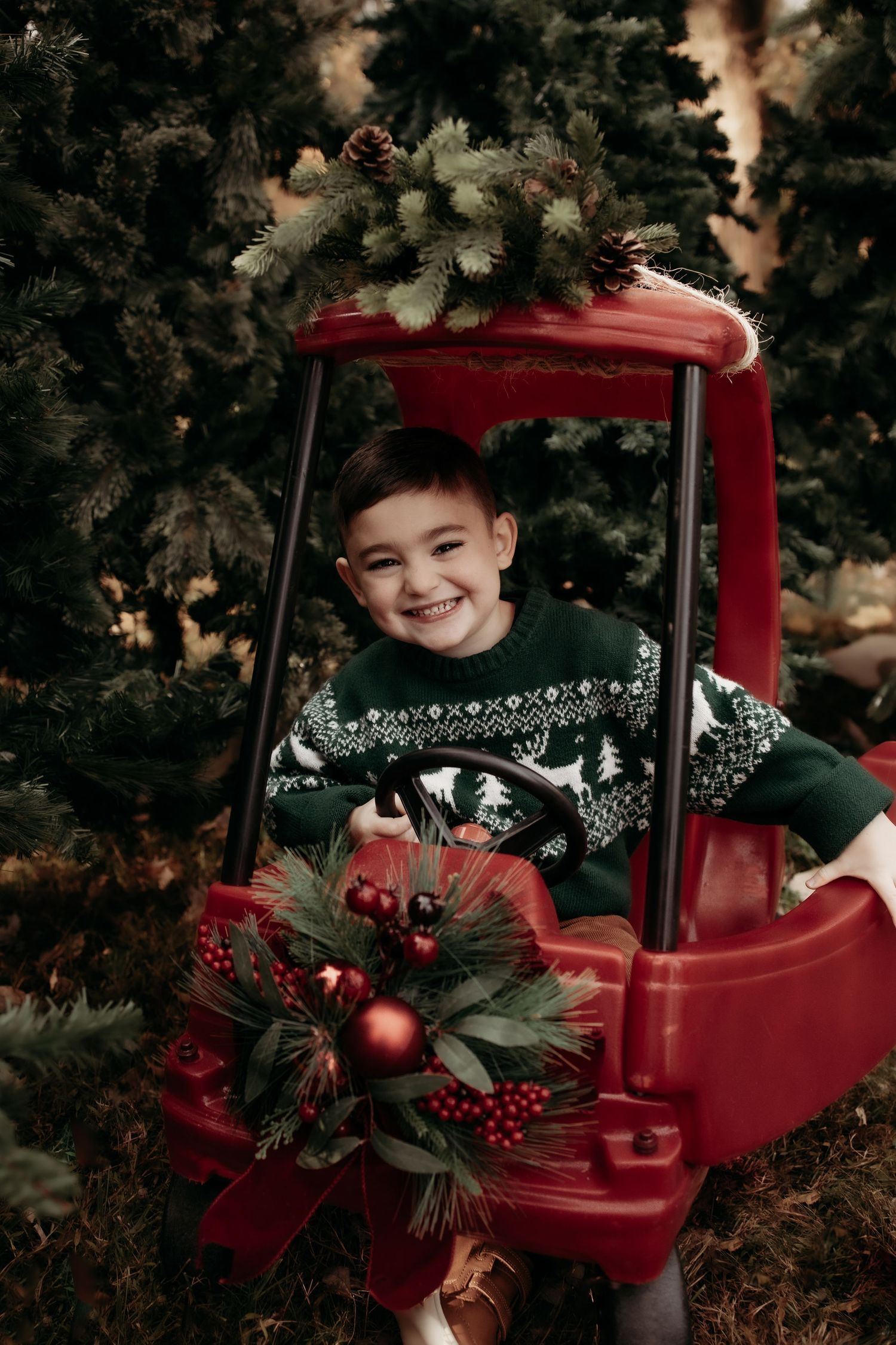 Smiling boy in green Nordic sweater sitting in red toy car surrounded by Christmas trees with holiday wreath decoration.