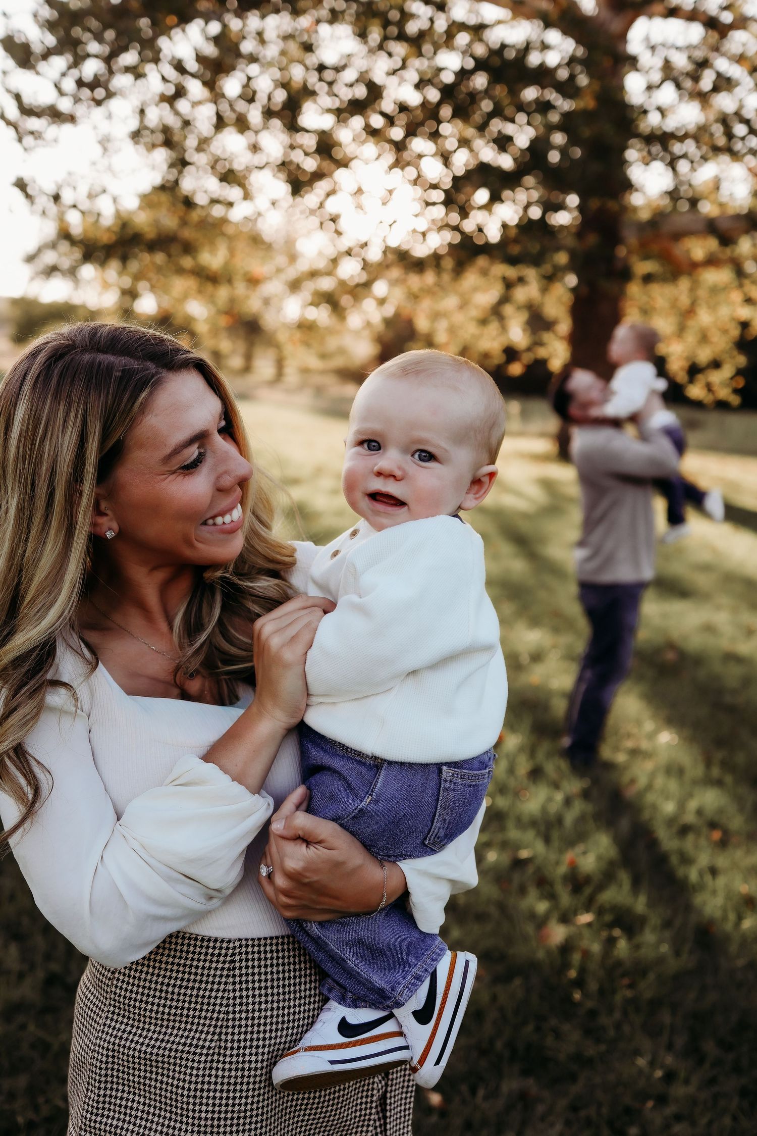 A smiling blonde mother holds her blue-eyed baby boy outdoors during a golden hour family portrait session.
