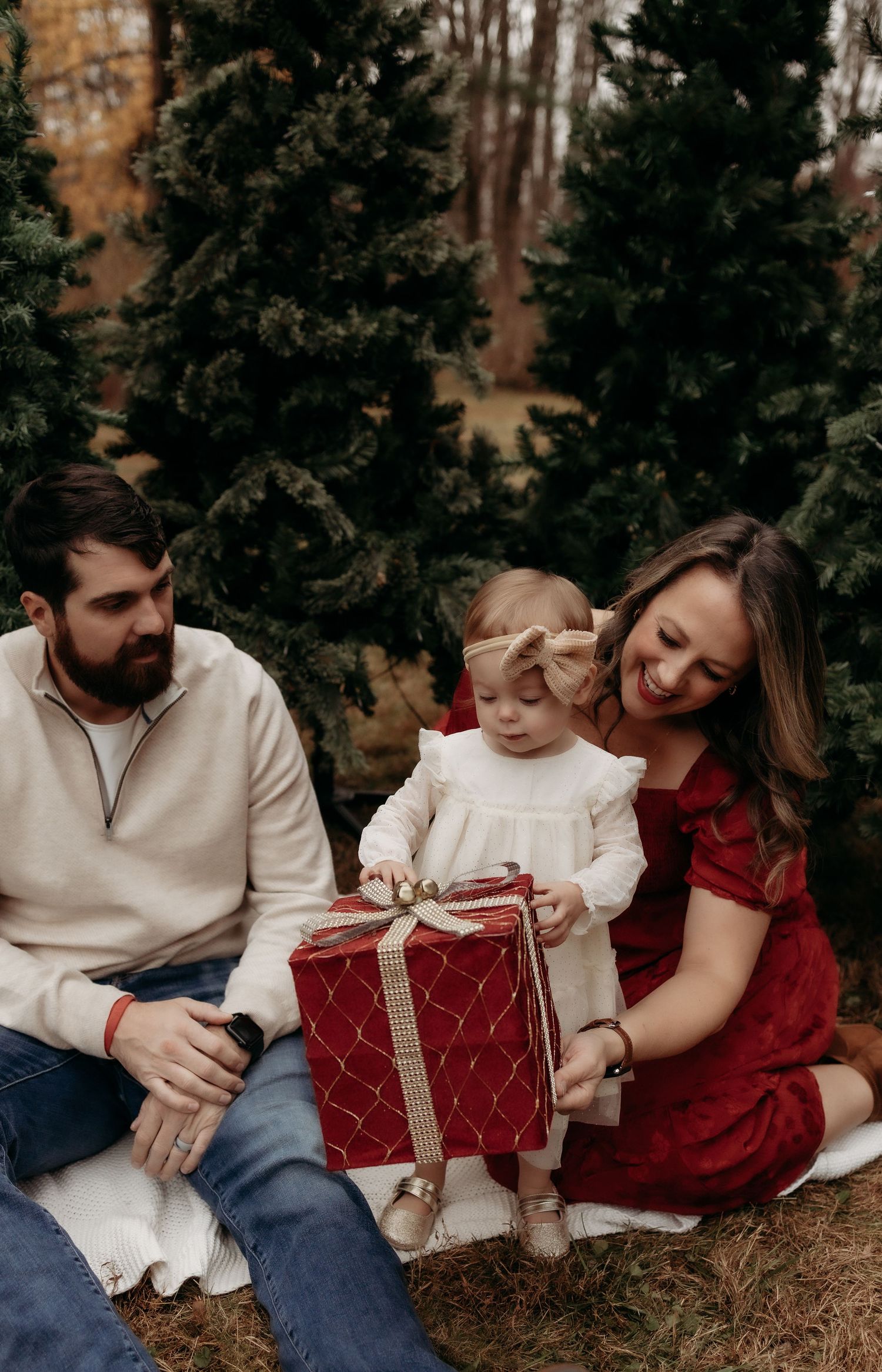 Family Christmas photo with parents and toddler opening a red gift box among evergreen trees outdoors.
