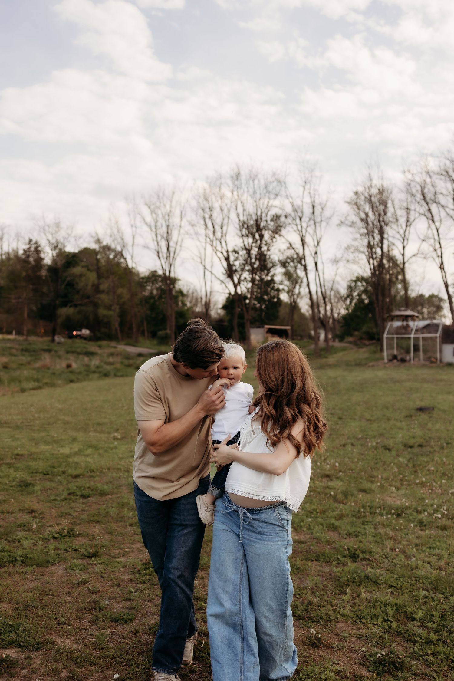 Young family with baby enjoying outdoor photo session in green field with trees in background.