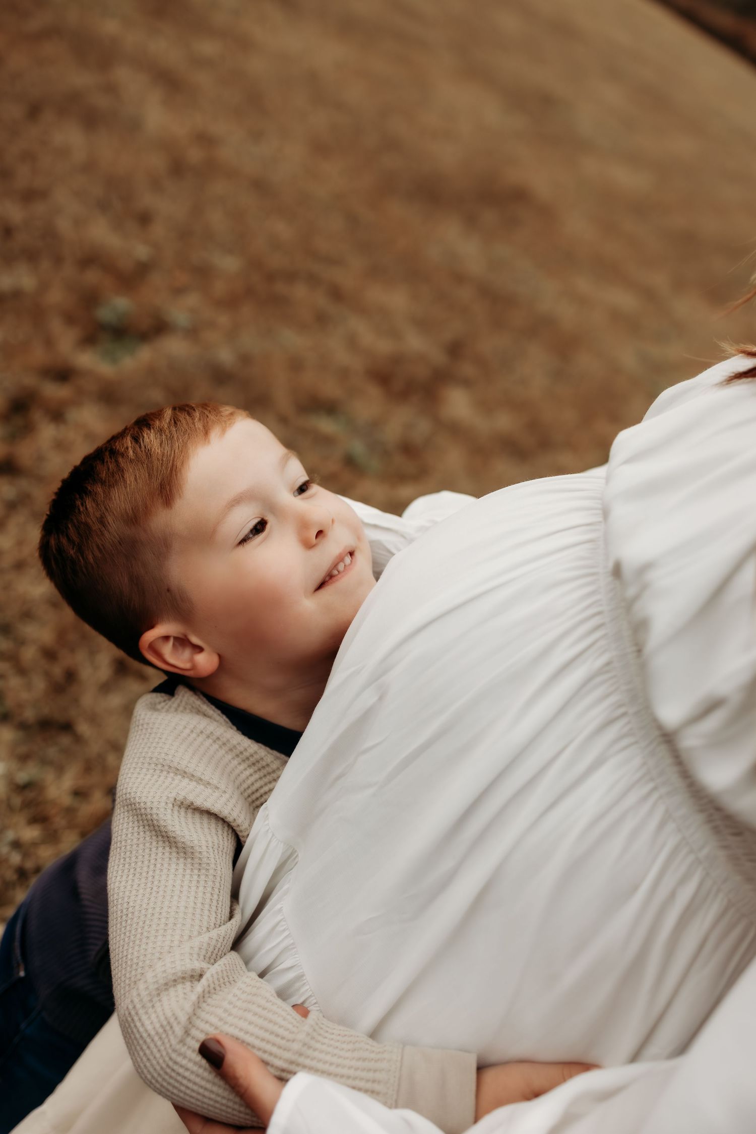 Smiling red-haired boy resting his head on a pregnant woman's baby bump outdoors.