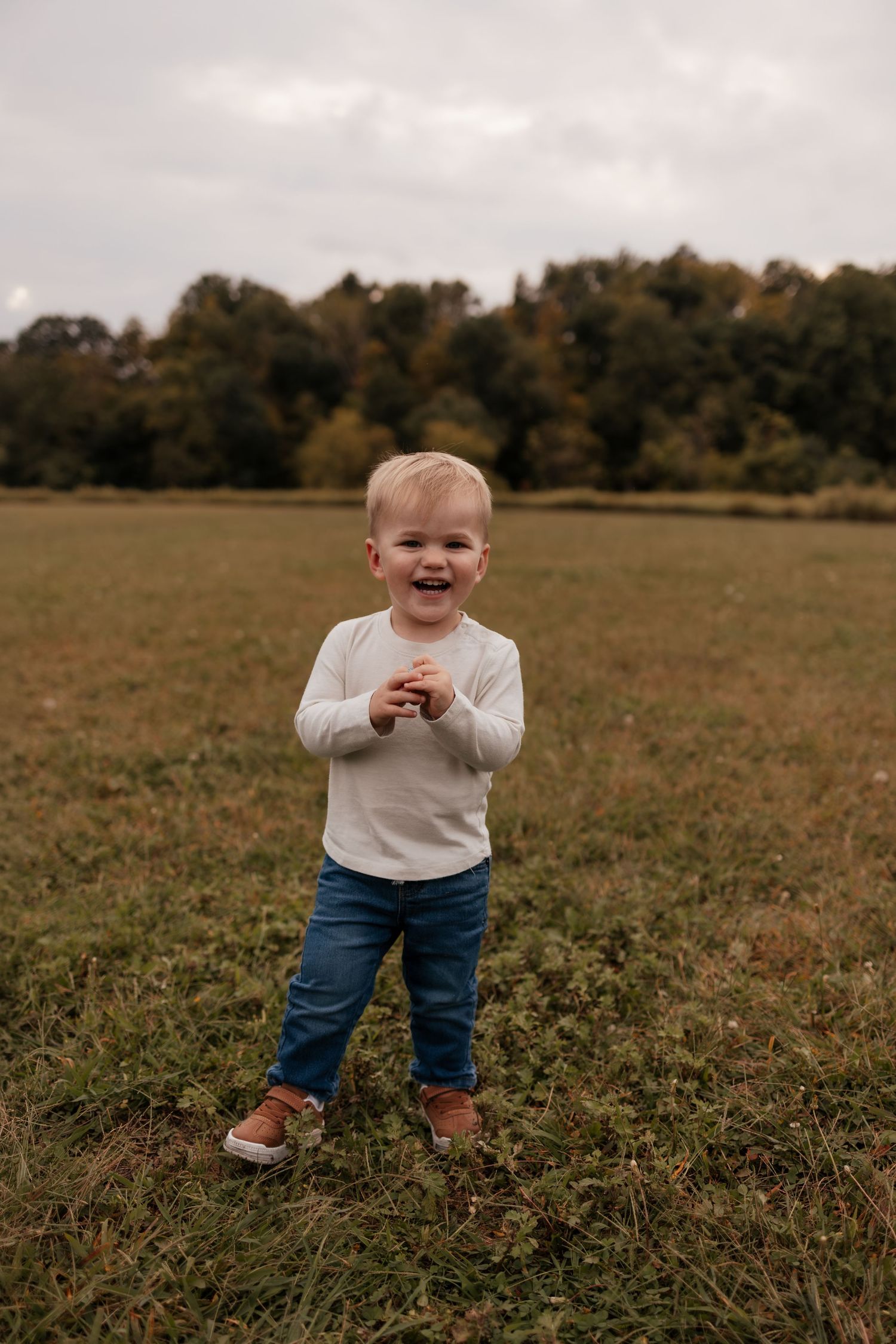 Smiling toddler boy in white shirt and jeans standing in green field with autumn trees in background.