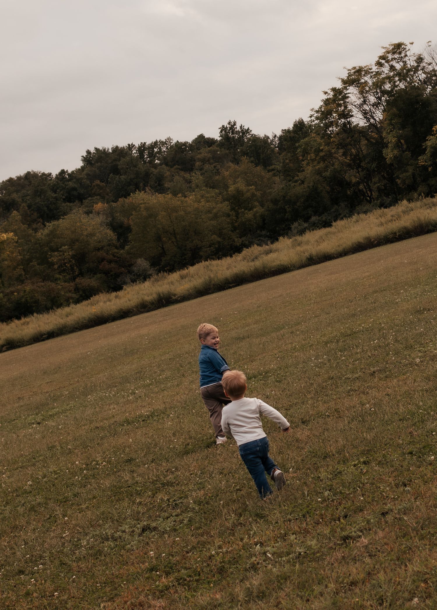Three young children running through a lush green field with autumn trees in the background on an overcast day.