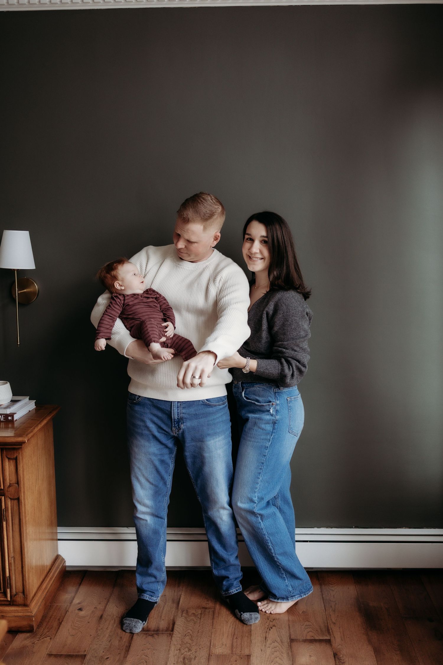 Young couple standing against dark wall holding newborn baby in striped outfit on hardwood floors.