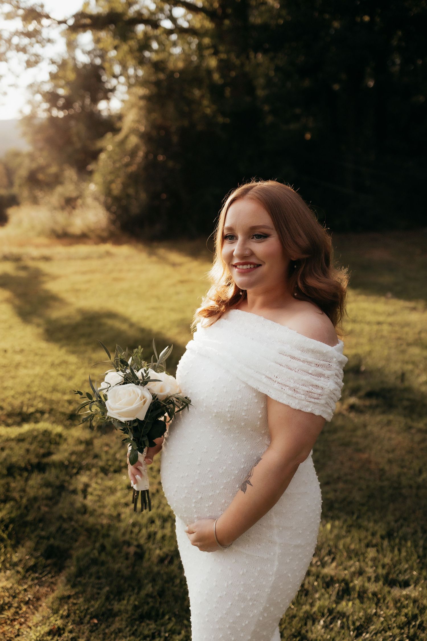 Pregnant bride in white off-shoulder gown holding bouquet of white roses, posing outdoors in golden hour light.