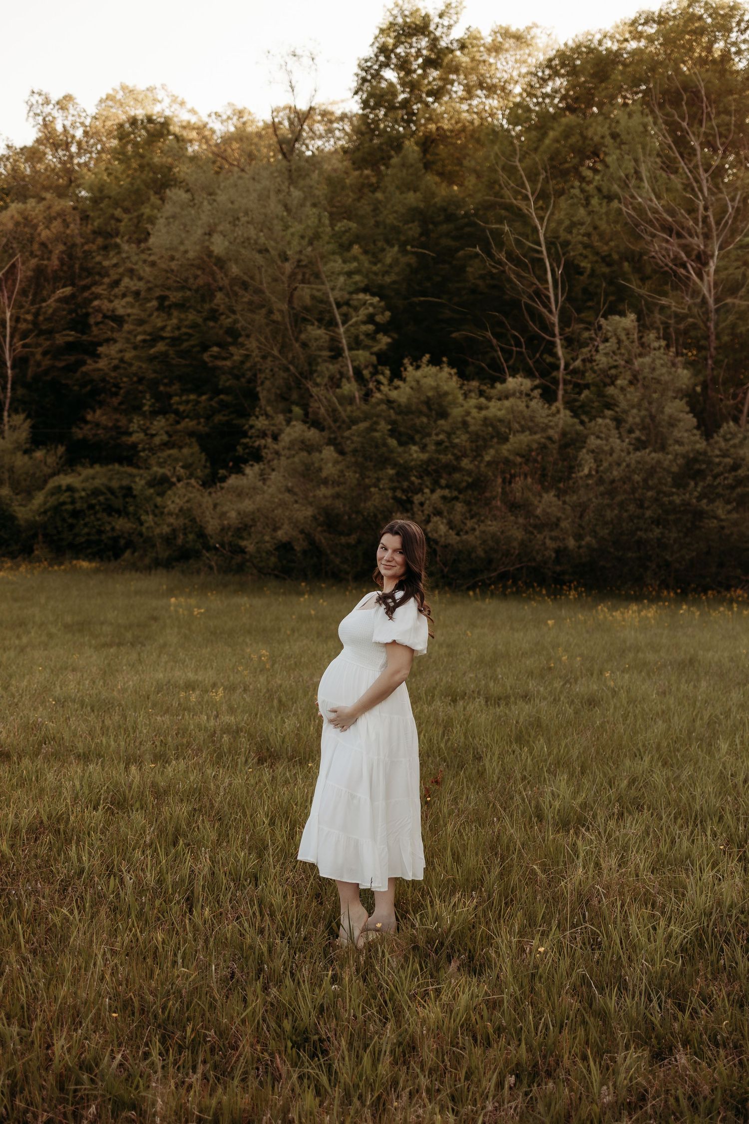 Pregnant woman in white off-shoulder dress standing in green meadow at sunset.
