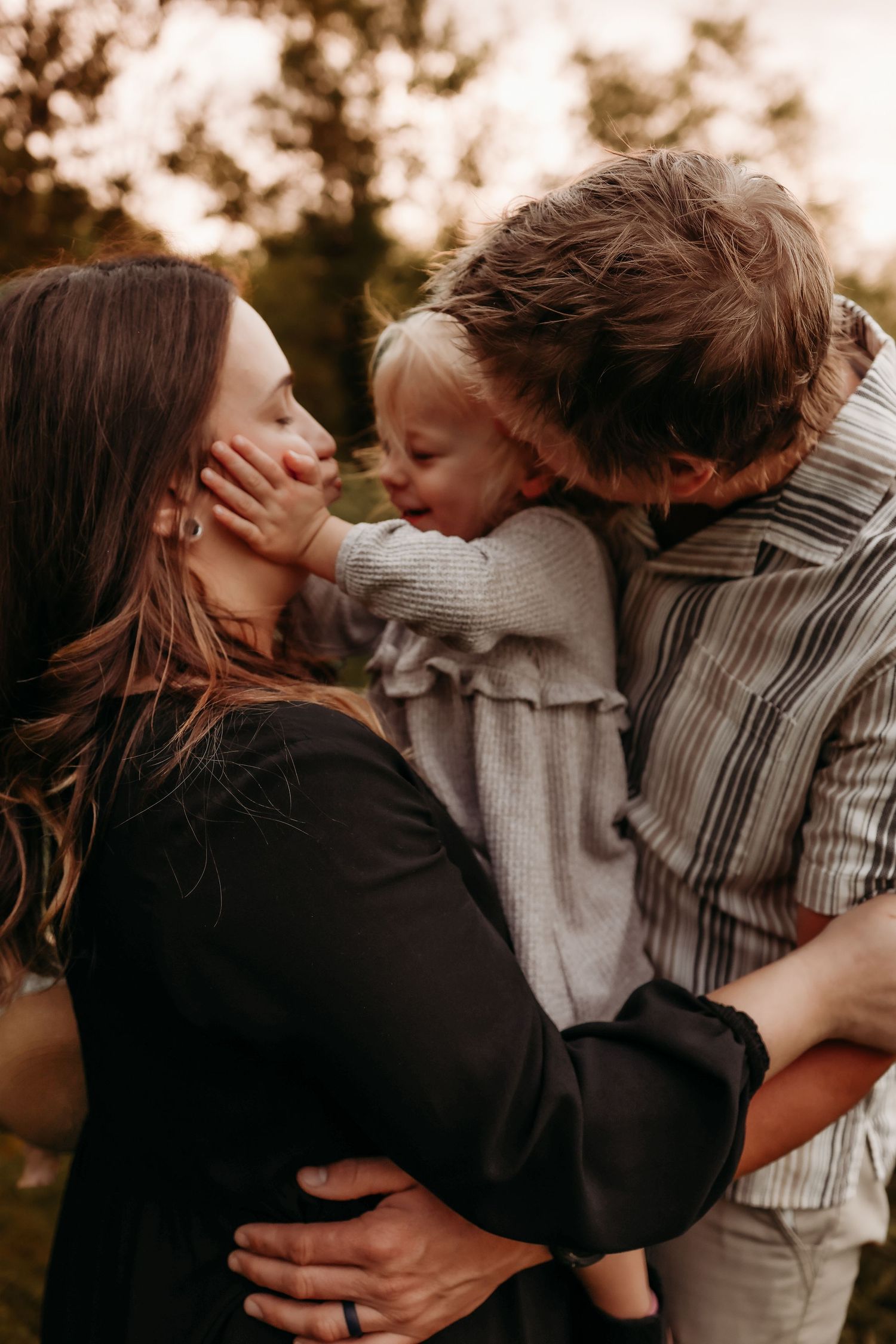 Parents share a loving moment with their toddler daughter between them in a warm outdoor setting.