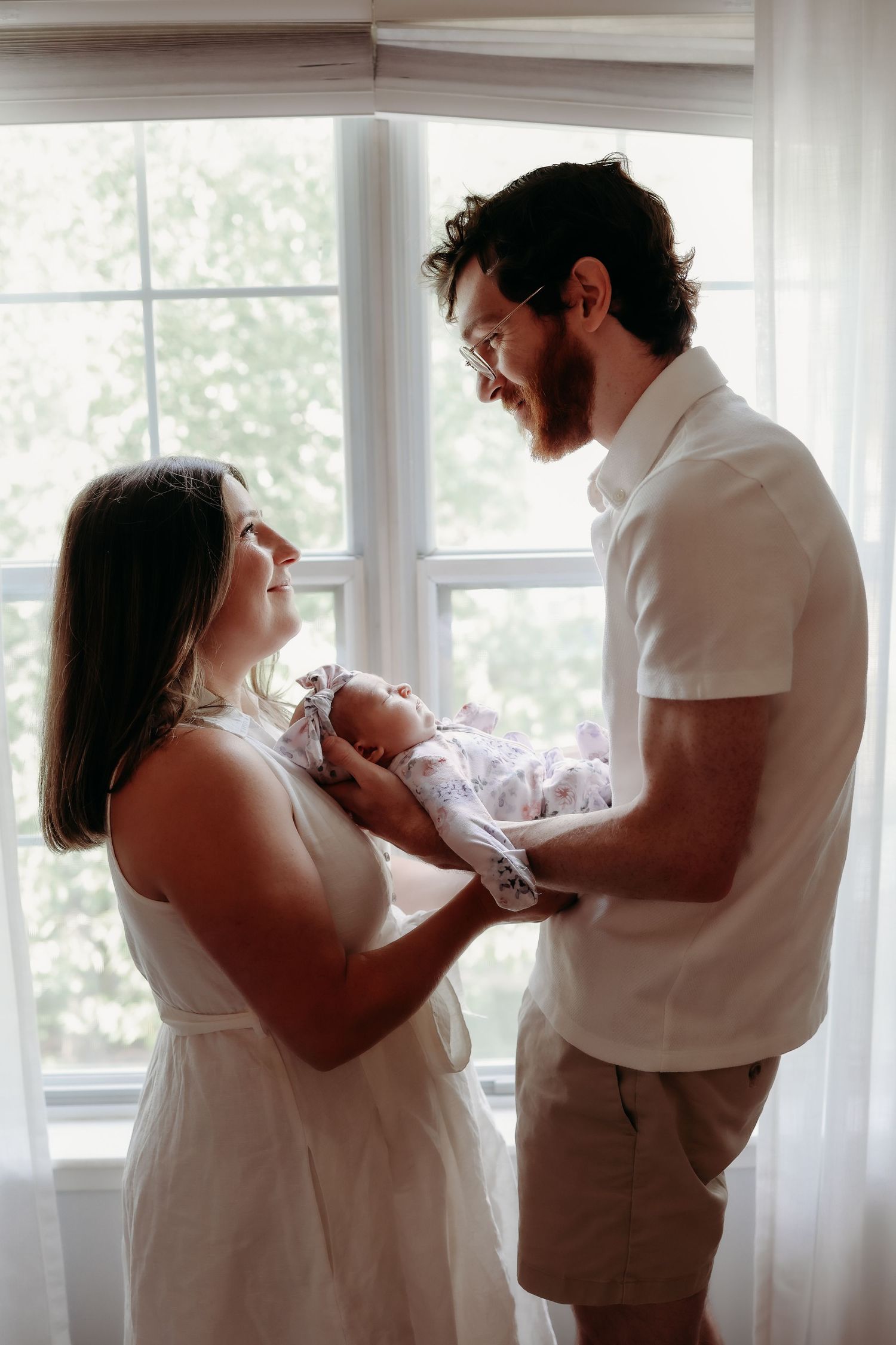 New parents holding newborn baby together in front of bright sunlit window at home.