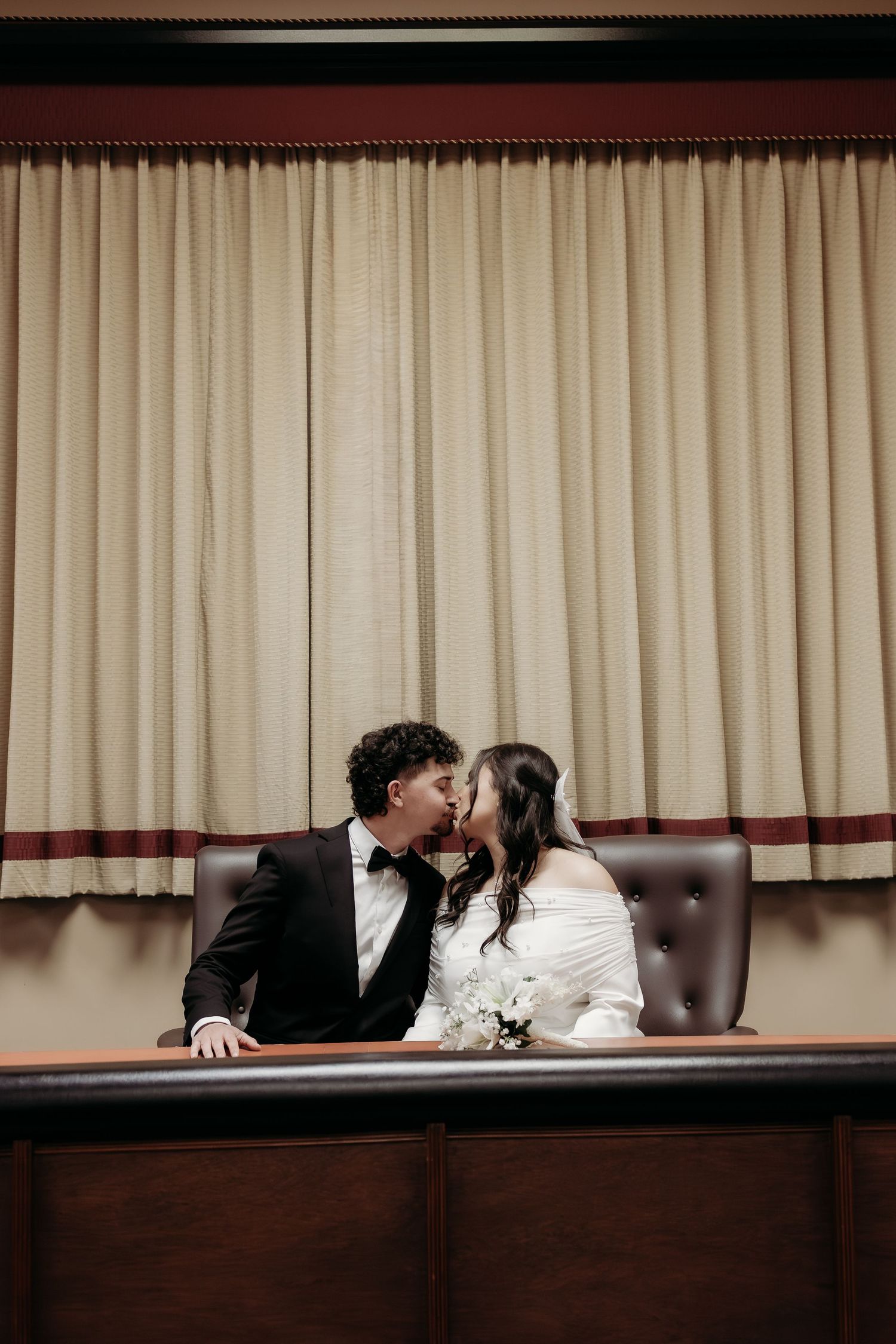 Newlyweds sharing a kiss while seated at a formal wooden table inside a courthouse chamber.
