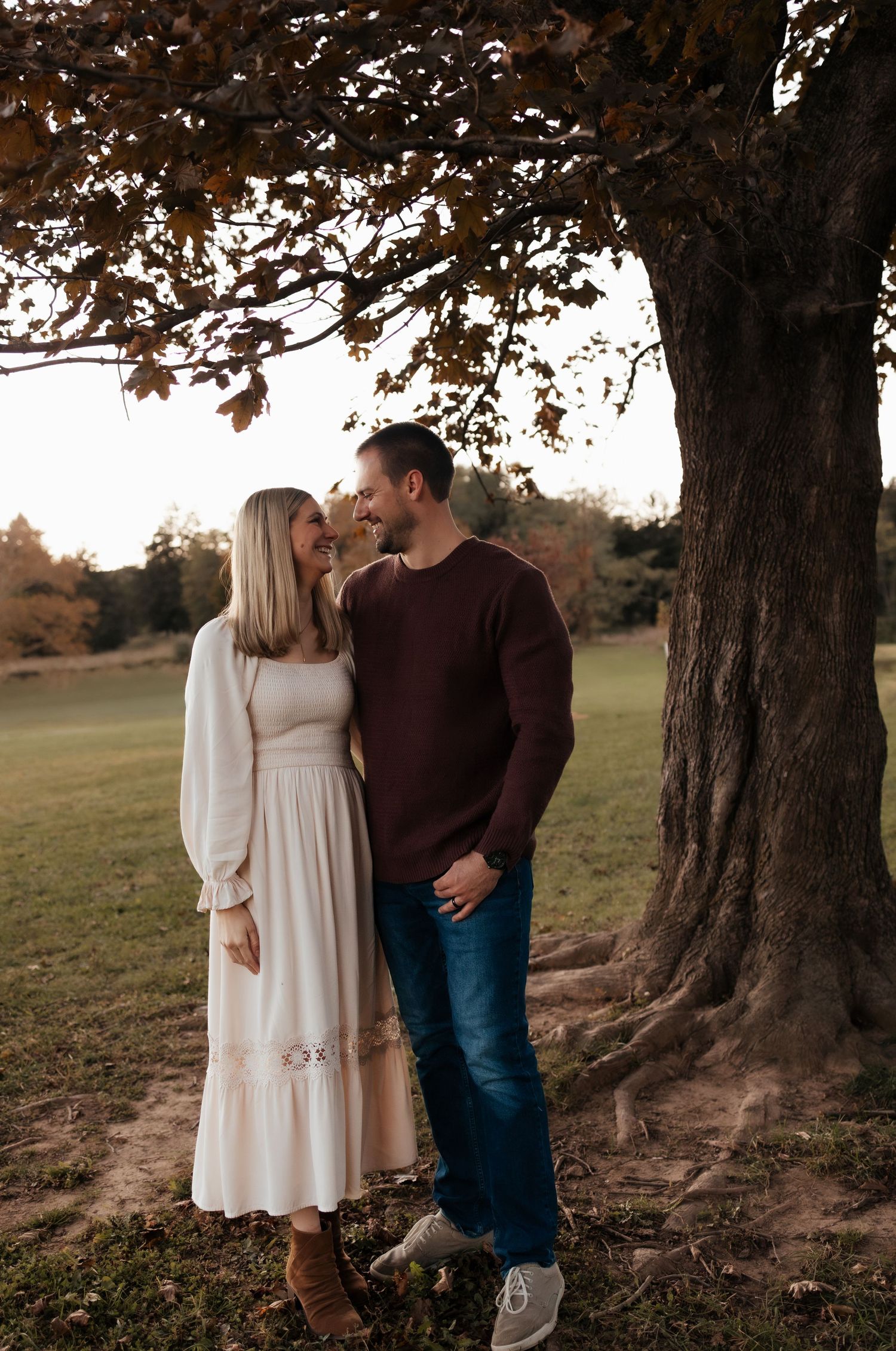 Smiling couple in a cream dress and maroon sweater embrace beneath a large autumn tree in a grassy park during golden hour.