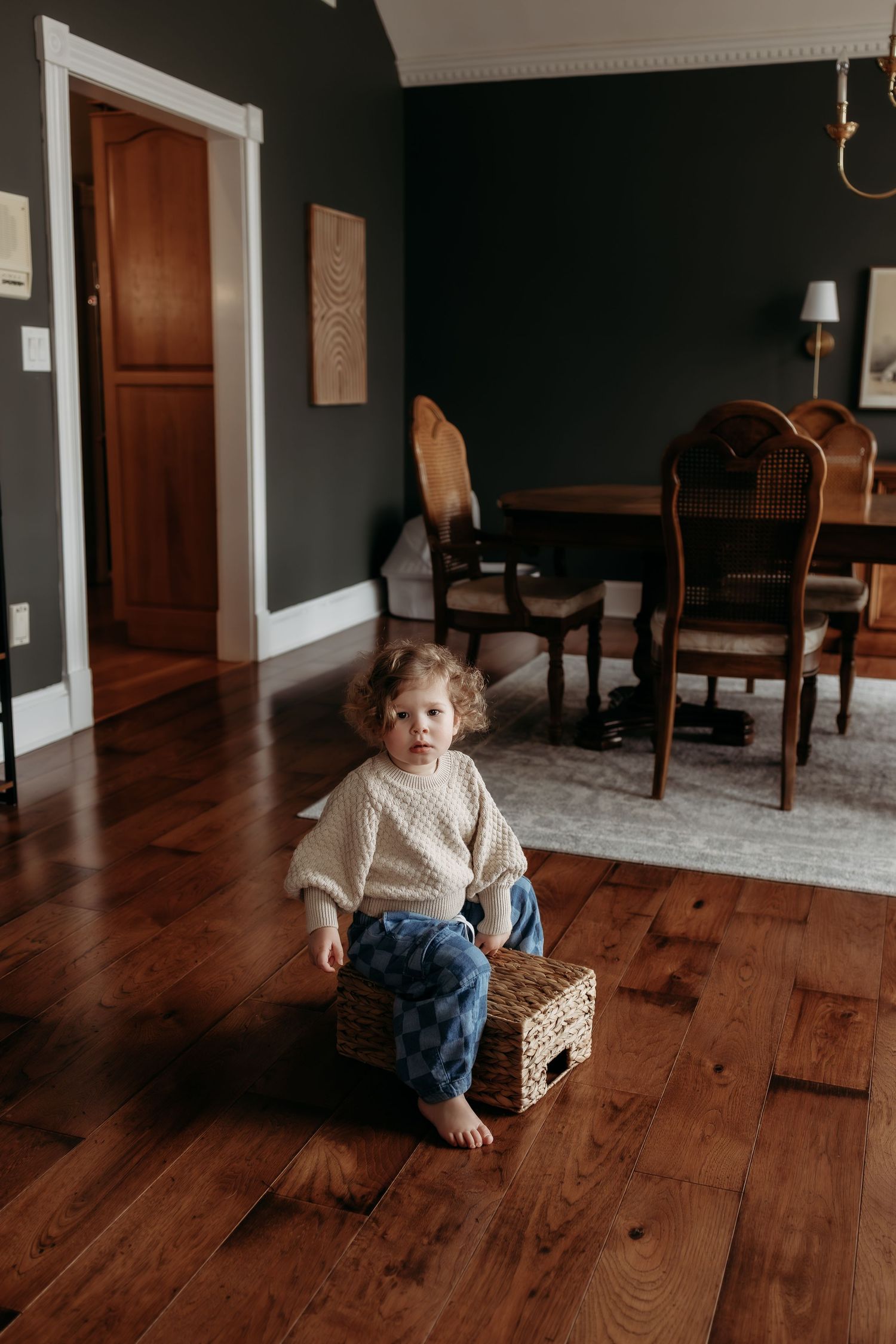 Toddler in cream sweater and plaid pants sitting on wicker stool on hardwood floor in dark-walled dining room.