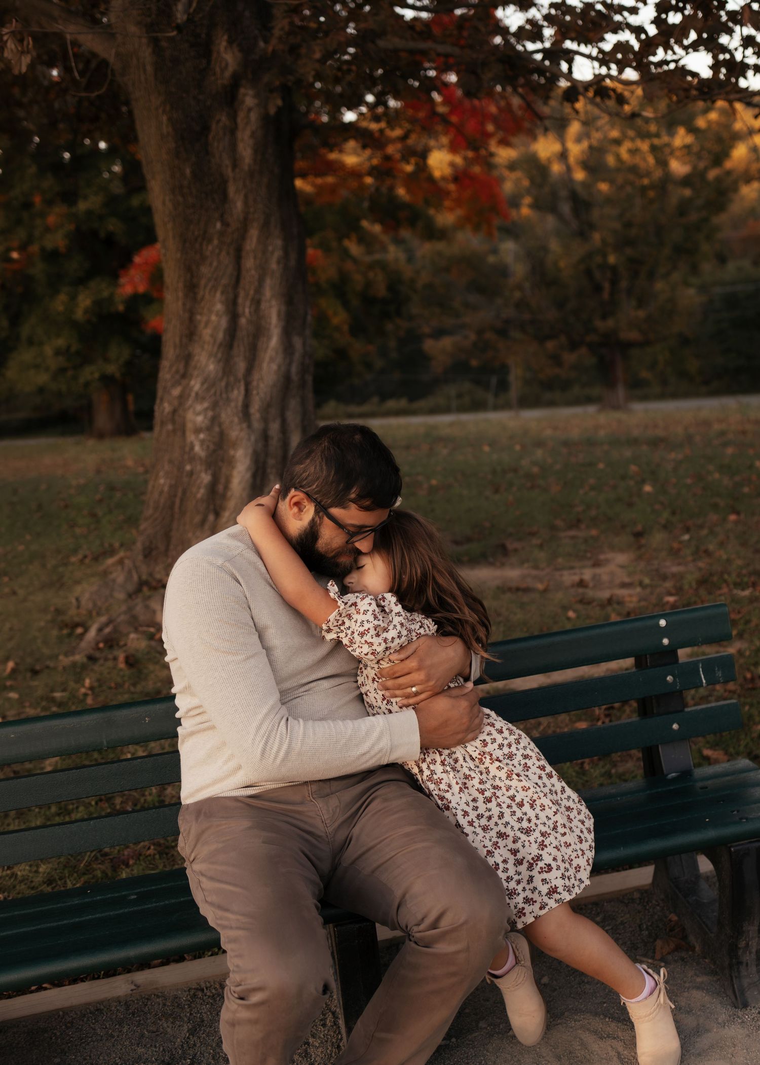 A bearded father in a cream sweater sits on a park bench embracing his young daughter in a floral dress beneath an autumn tree.
